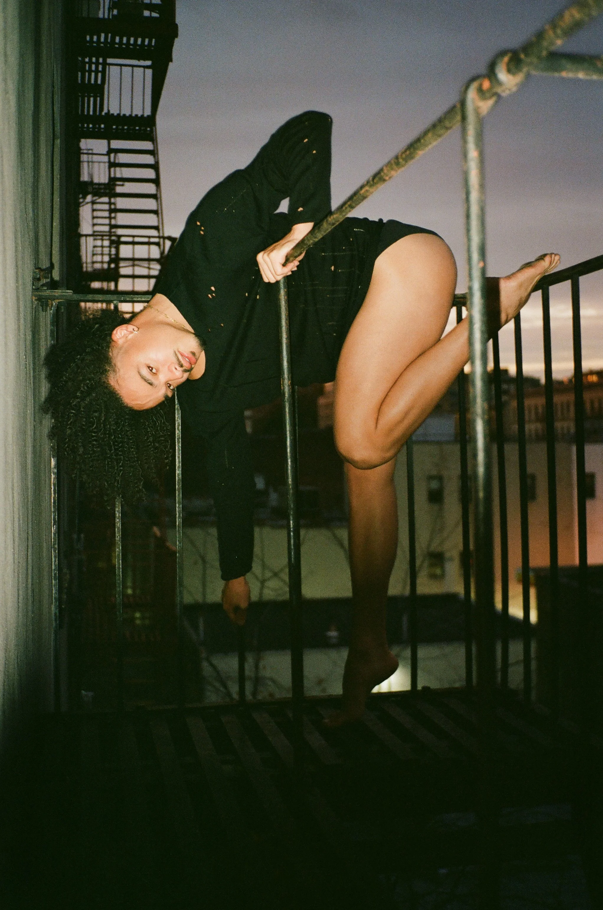 A woman with curly hair hanging upside-down on an outdoor fire escape at dusk, holding onto the railing with both hands and smiling at the camera.