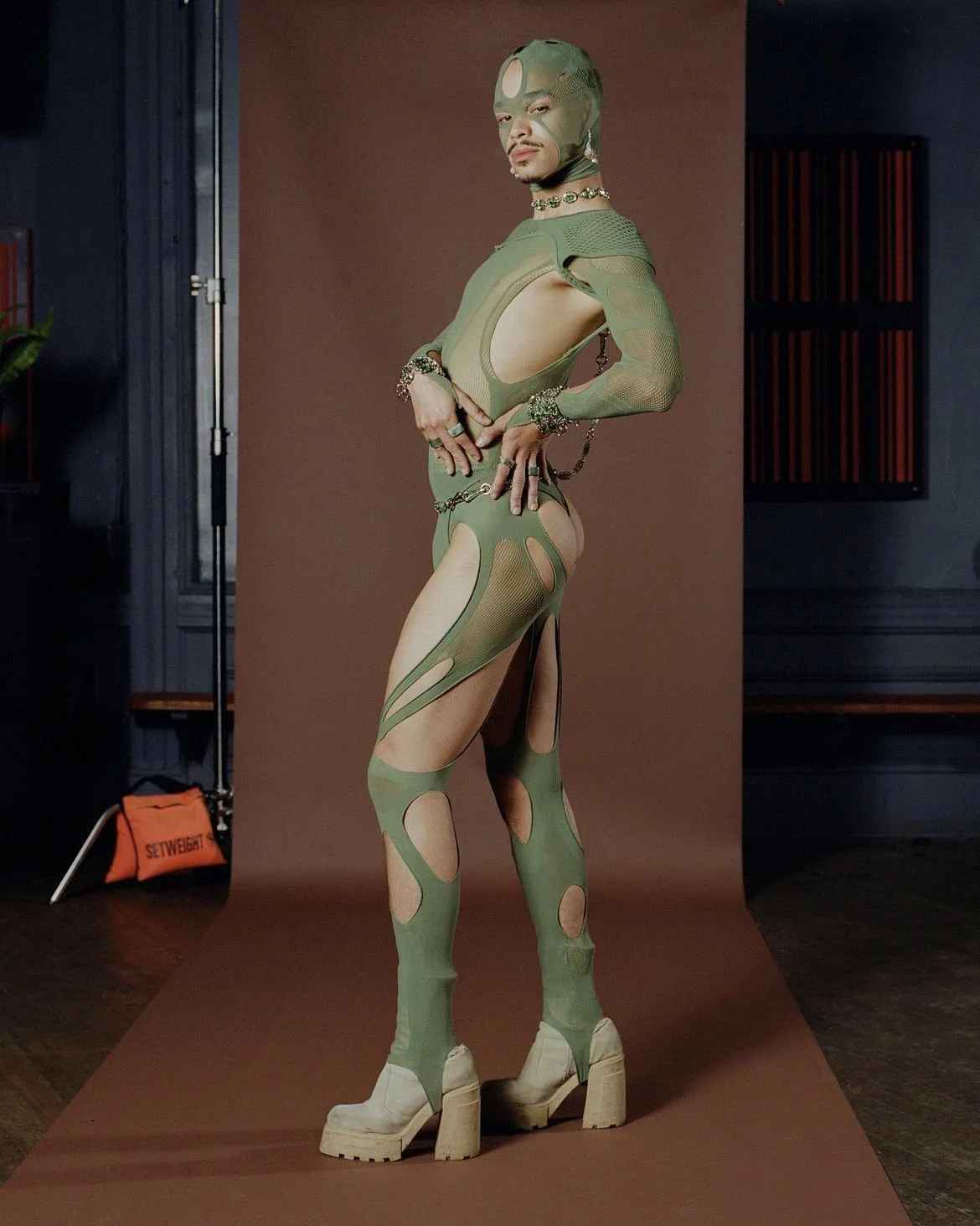 A model in a futuristic, green cut-out outfit with matching headgear, standing on a brown backdrop in a photography studio.