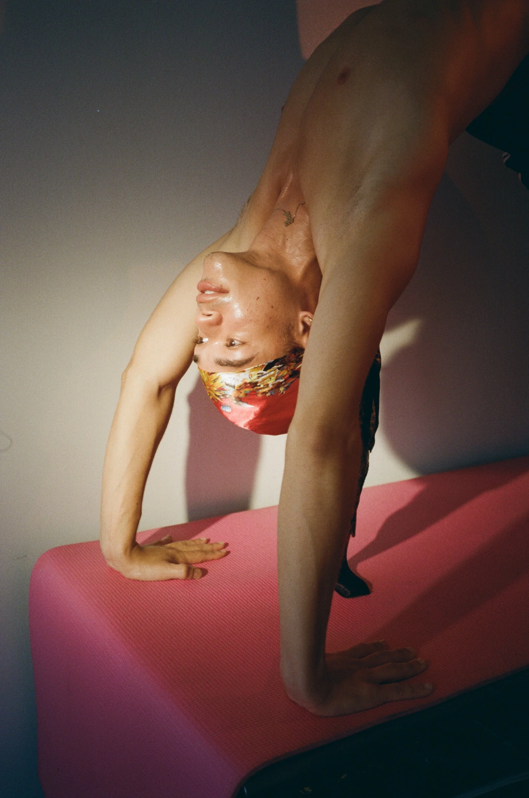 A woman practicing yoga in a downward dog pose on a pink yoga mat, wearing a colorful headscarf, indoors with soft lighting.
