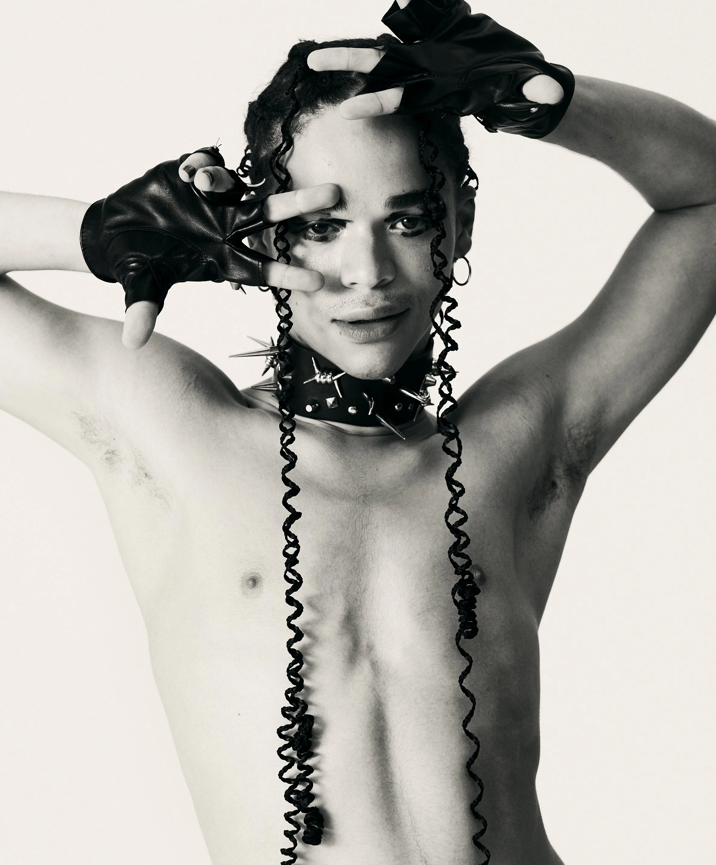 A shirtless person with long, curly hair styled in two long braids, wearing spiked accessories and fingerless gloves, making a peace sign near their face.