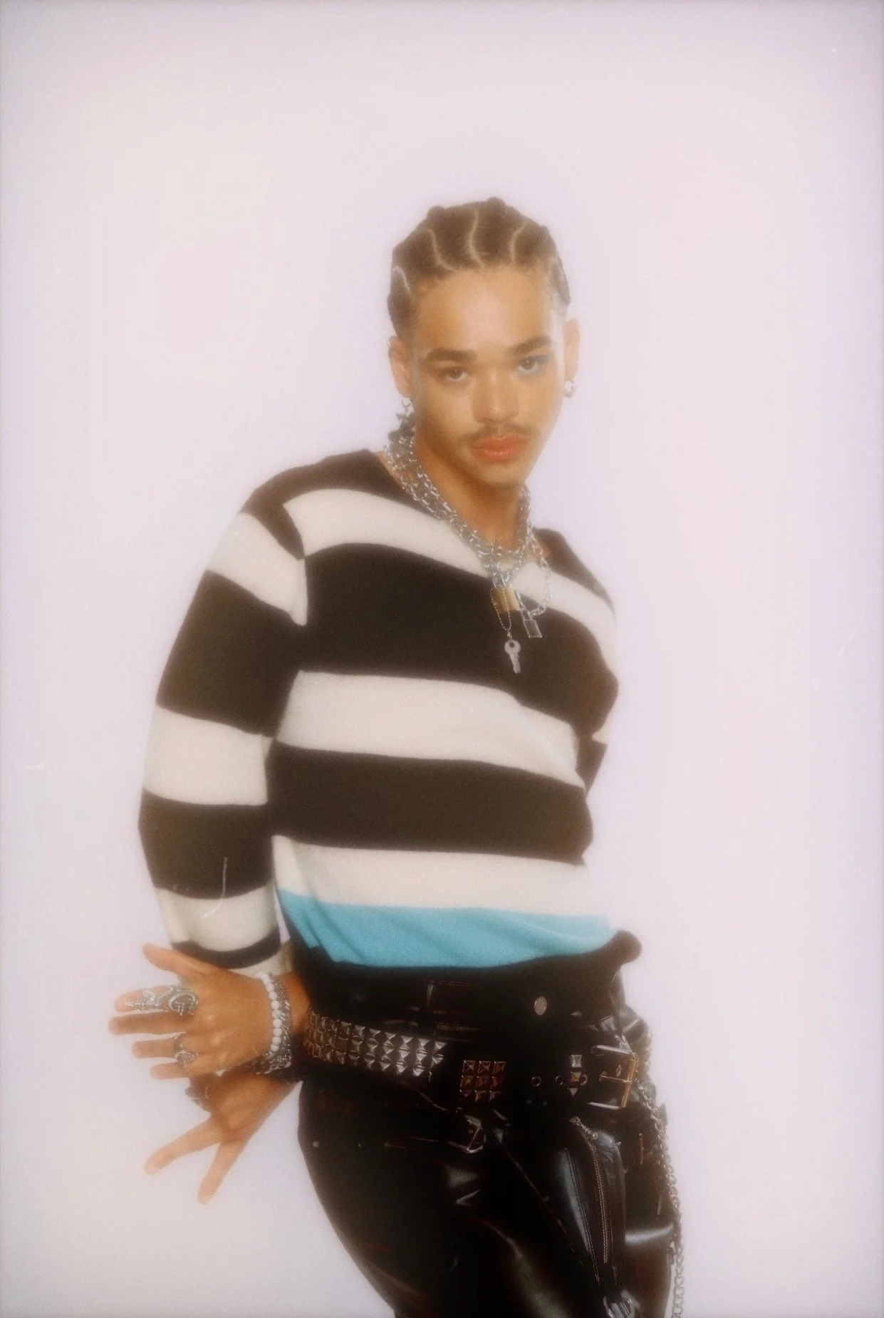 A person with braided hair wearing a striped sweater, multiple jewelry, and black leather pants posing against a plain background.