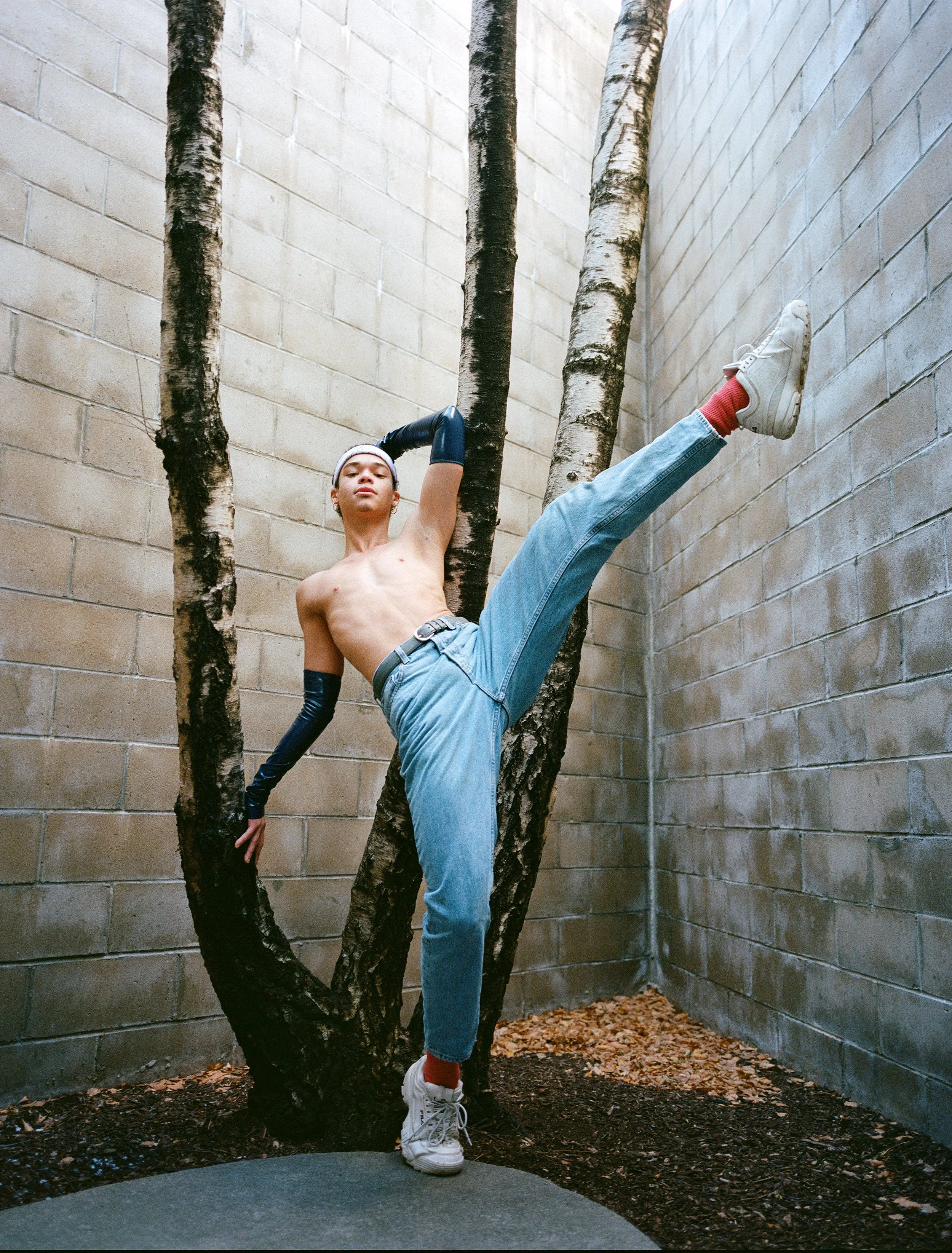 A shirtless young man wearing jeans, white sneakers, red socks, black gloves, and a headband strikes a dance pose in an outdoor corner with bare-brick walls and a small tree.