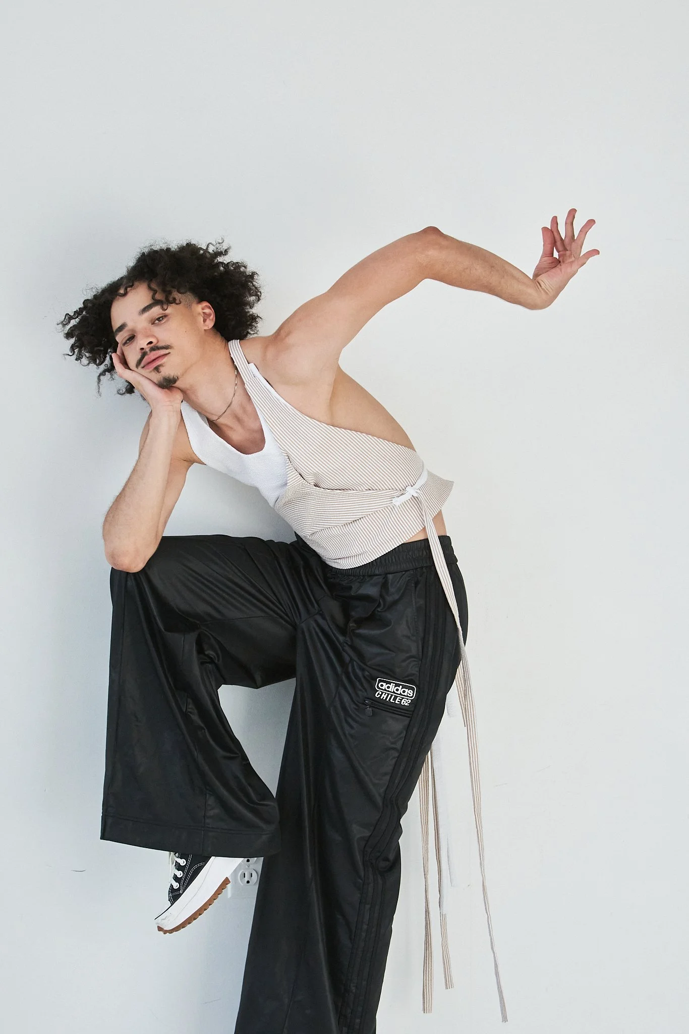 A man with curly hair and facial hair, wearing a sleeveless top and black Adidas track pants, is posing against a plain white wall, balancing on one leg with his hand resting on his face and the other arm extended.