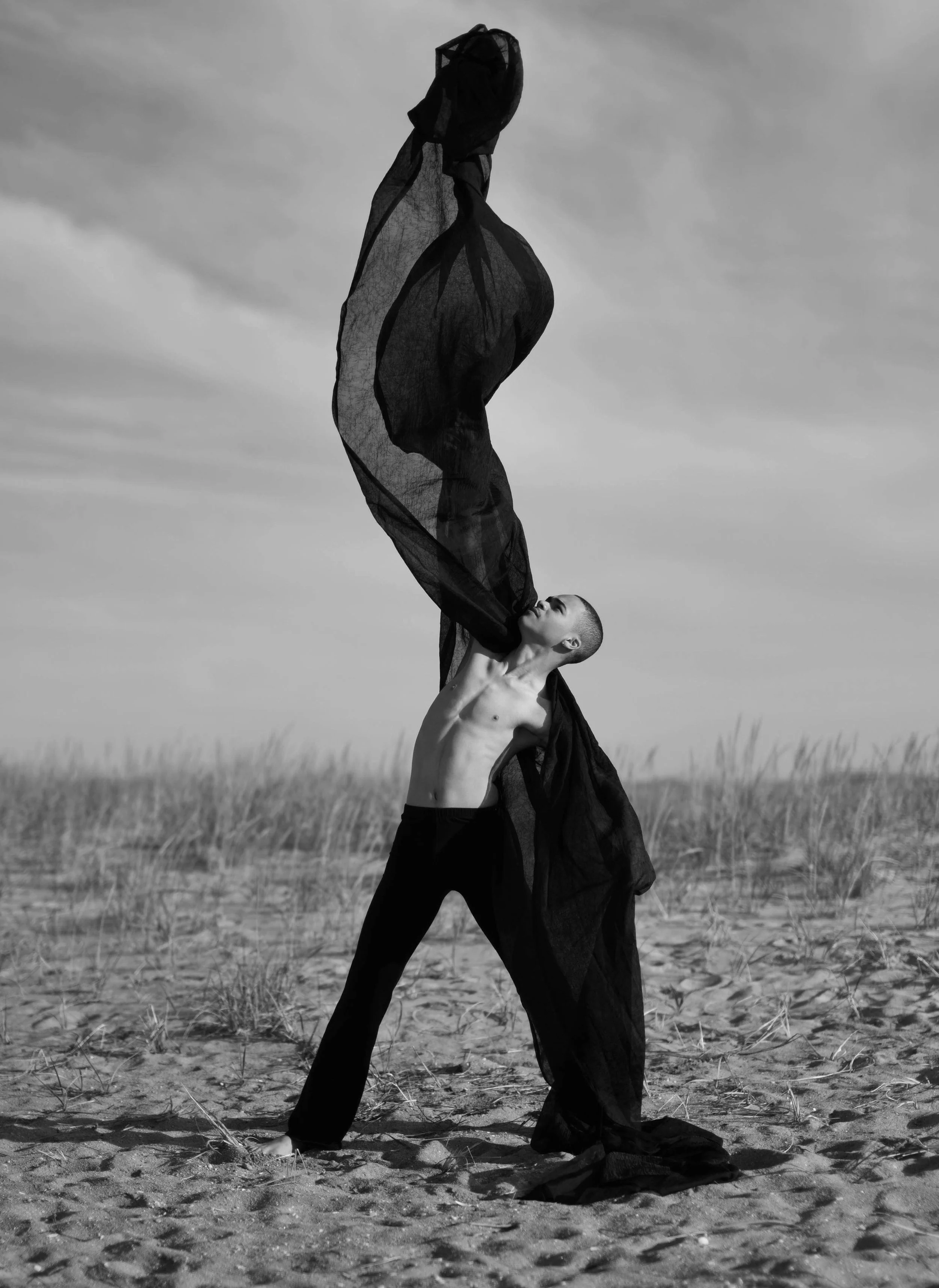 A black and white photograph of a shirtless person with a shaved head standing on a sandy beach. They are holding a large, flowing piece of black fabric above their head with one arm extended upward. The person appears to be in a dance pose, looking 