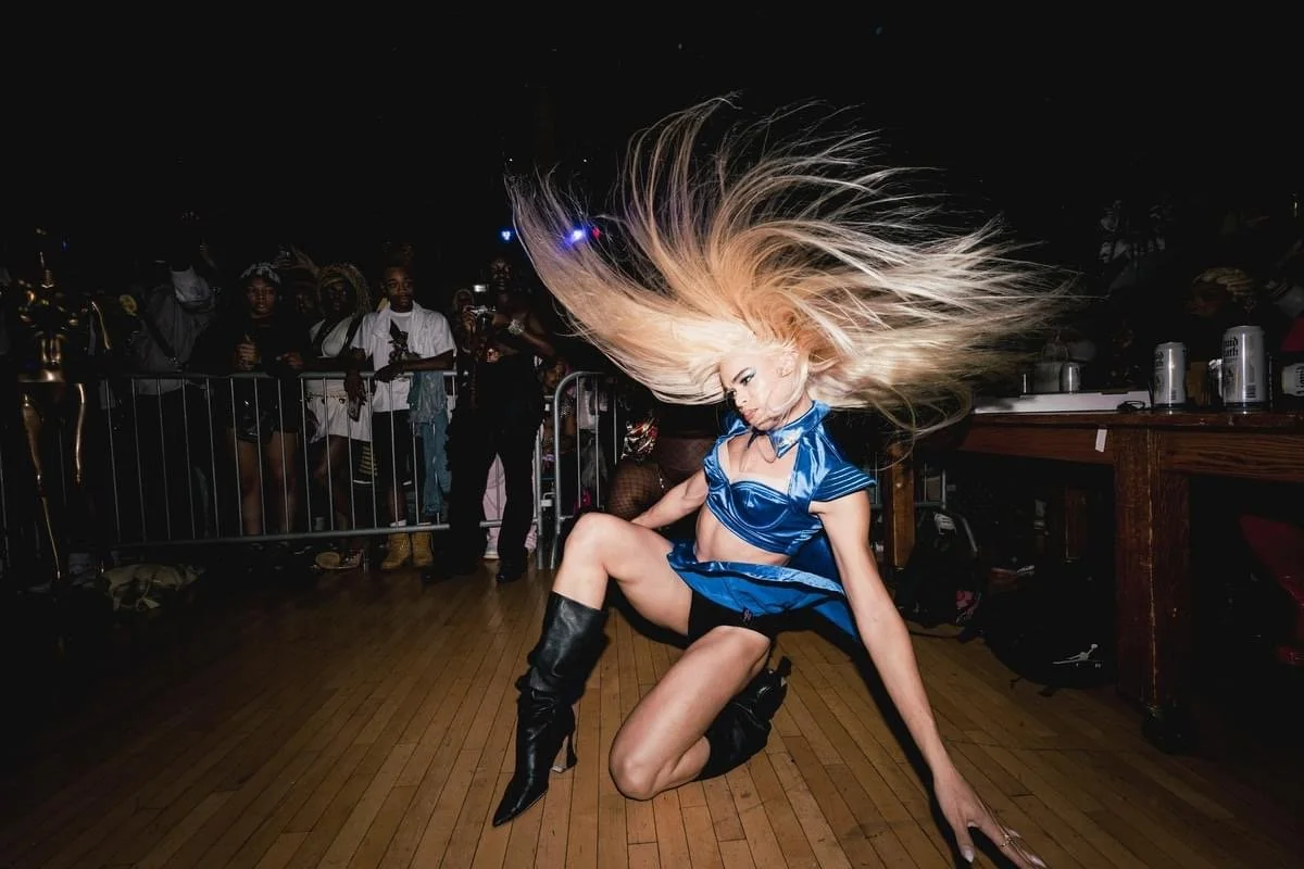 A female dancer with long blonde hair styled upward, wearing a blue satin crop top, matching skirt, and black knee-high boots, dancing on a wooden floor at a lively event with people watching in the background.