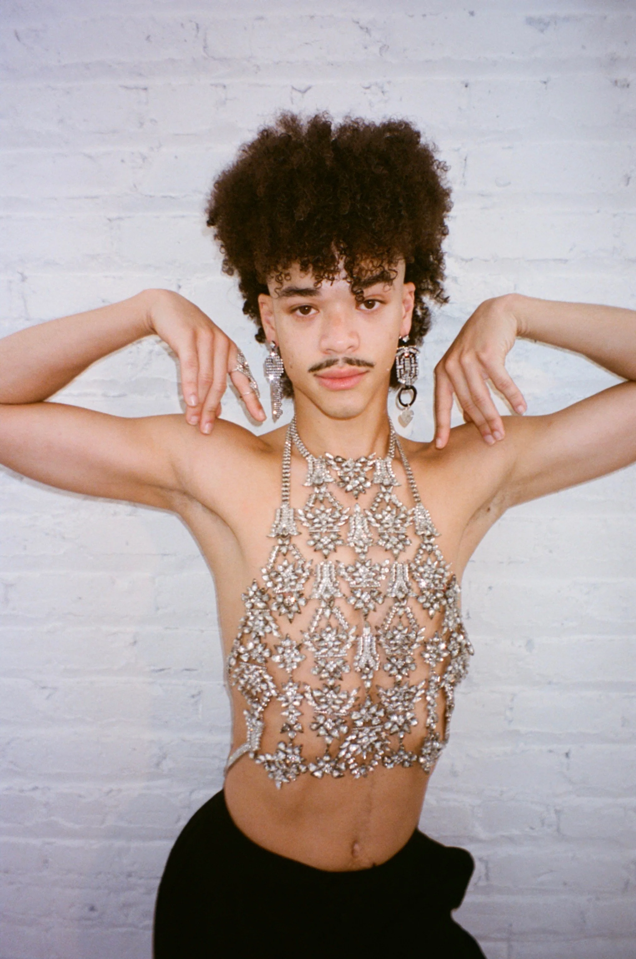 Person wearing large, crystal jewelry with curly hair posed against a white brick wall.