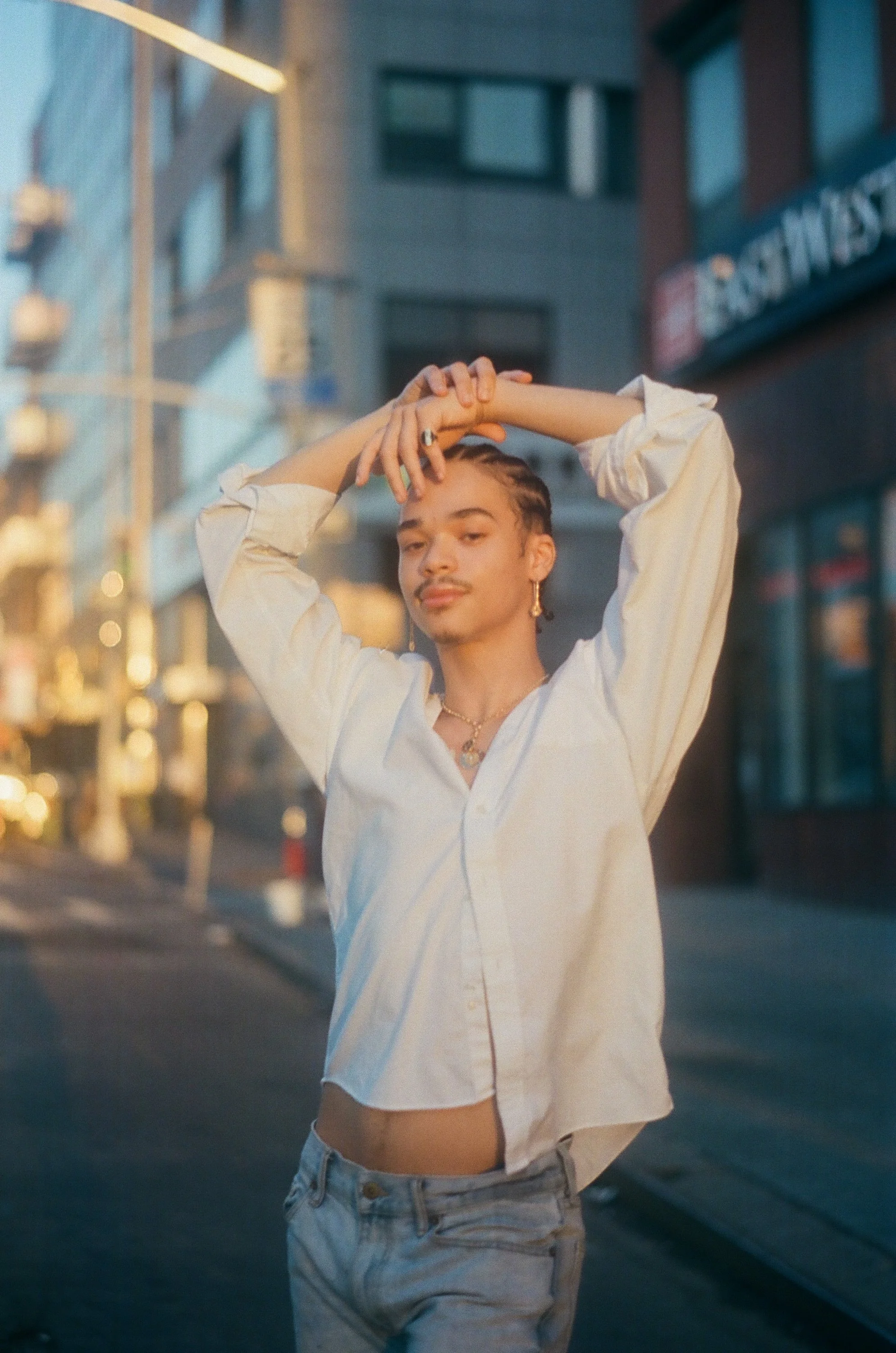 A young person with light skin, short braided hair, and jewelry, standing on a city street at sunset, wearing a white shirt and jeans, looking at the camera with a relaxed expression.