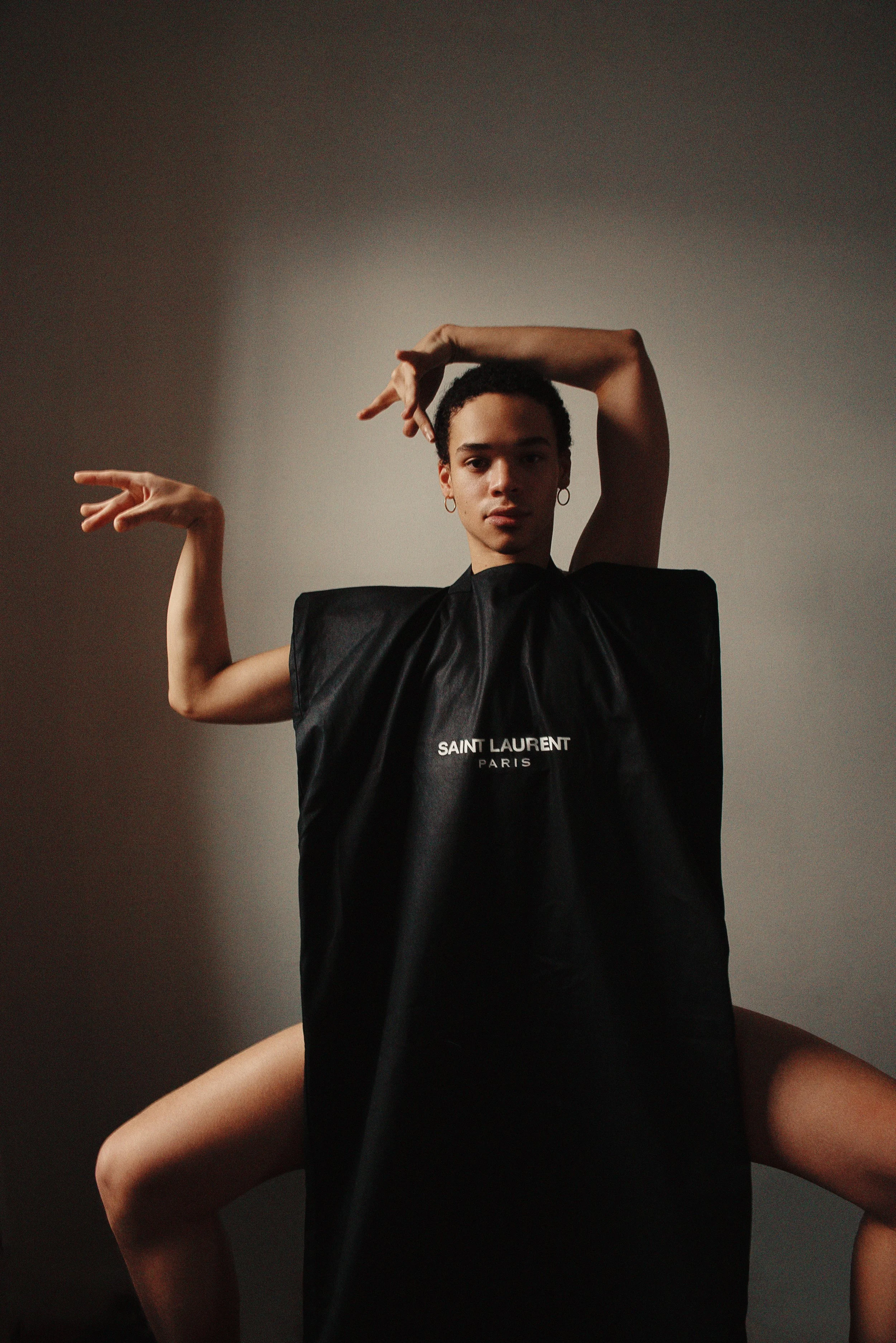 A person wearing a black Saint Laurent Paris garment poses with arms bent and hands in a dance-like gesture against a neutral background.