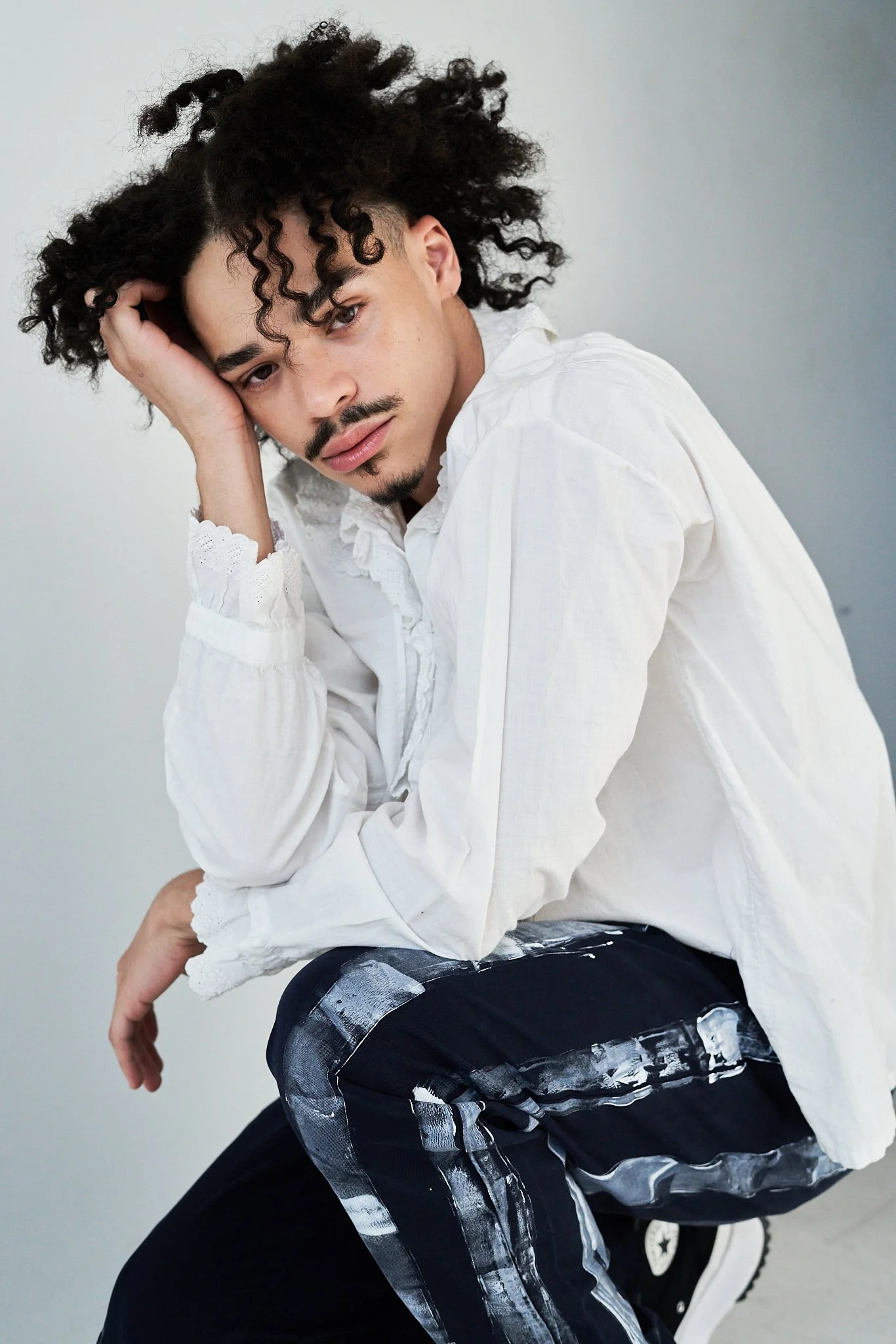 A young man with curly dark hair and a mustache, wearing a white shirt, is crouching down with one hand resting on his head, looking at the camera against a plain gray background.