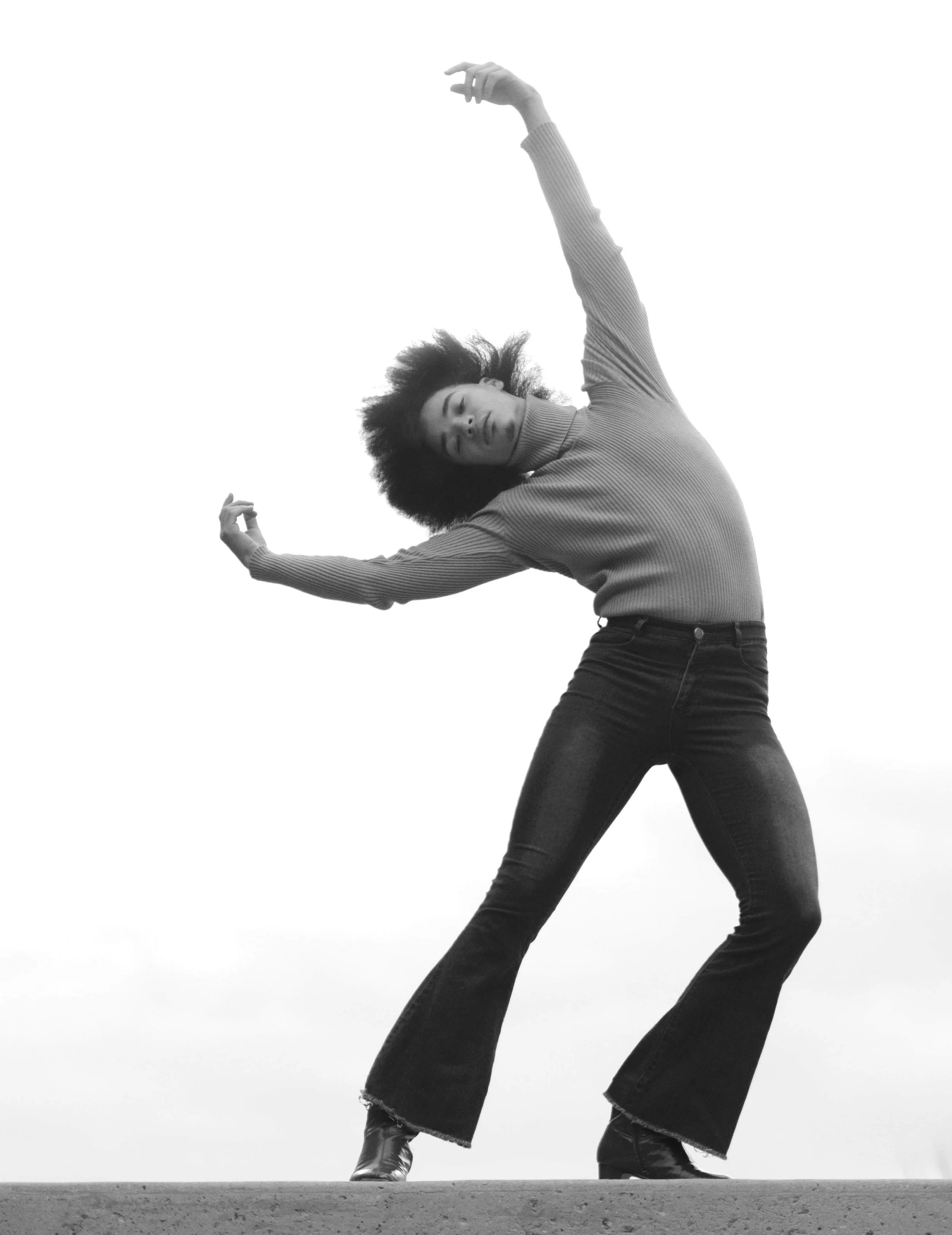 A woman with curly hair wearing a turtleneck sweater and flared jeans, standing on a flat surface, stretching her arms above her head, against a plain, light background.