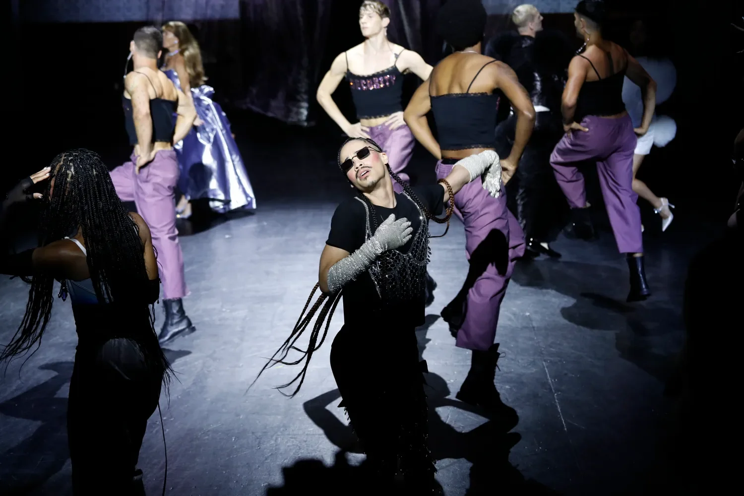 A group of diverse performers on stage, dressed in colorful and eclectic costumes, mid-performance with one prominent performer in the foreground wearing sunglasses and dramatic gloves, striking a pose.