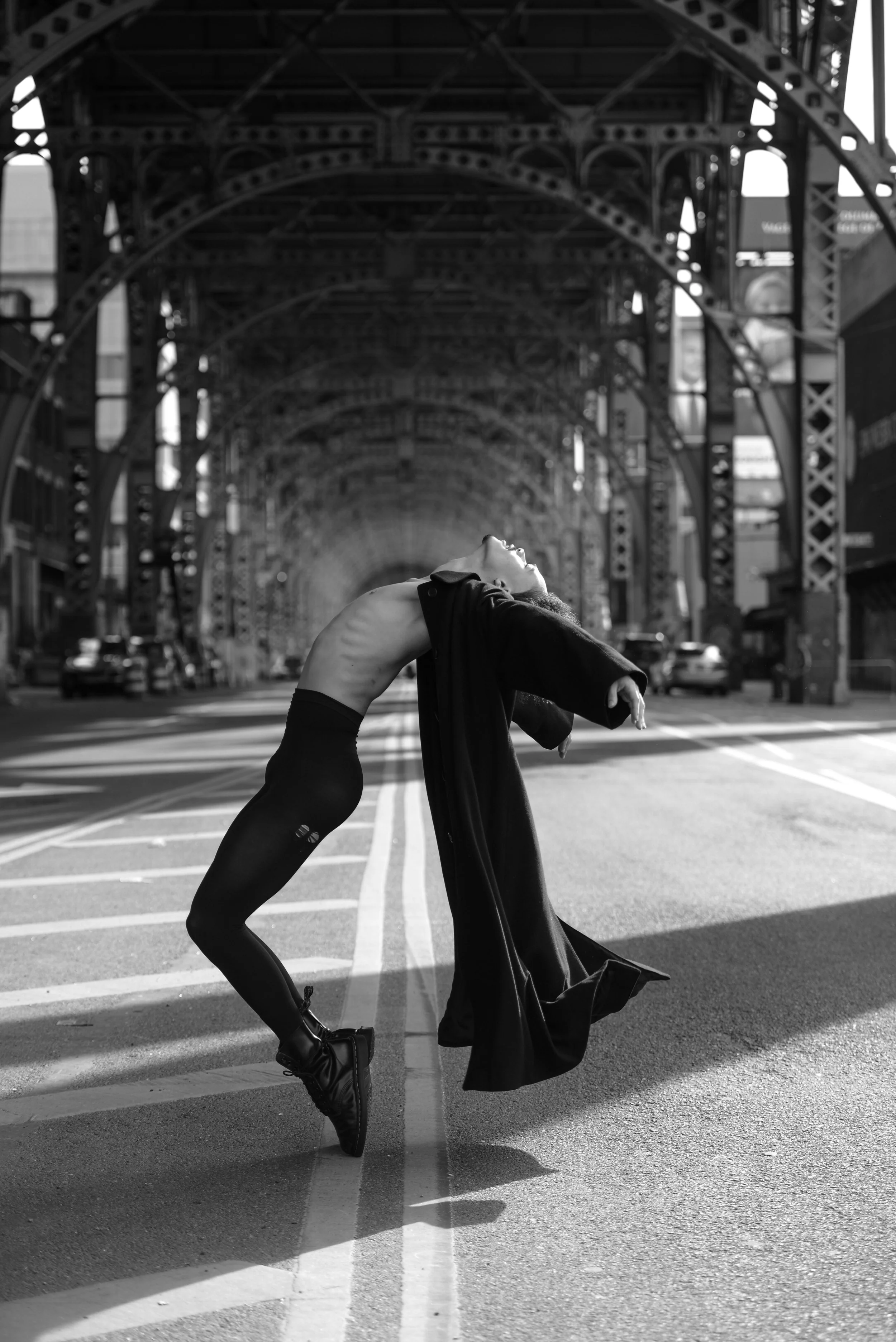 A person with a shirt draped over their shoulders, wearing tights and shoes, bending backwards in a dance pose on an empty city street under metal bridge arches, with cars and buildings in the background.