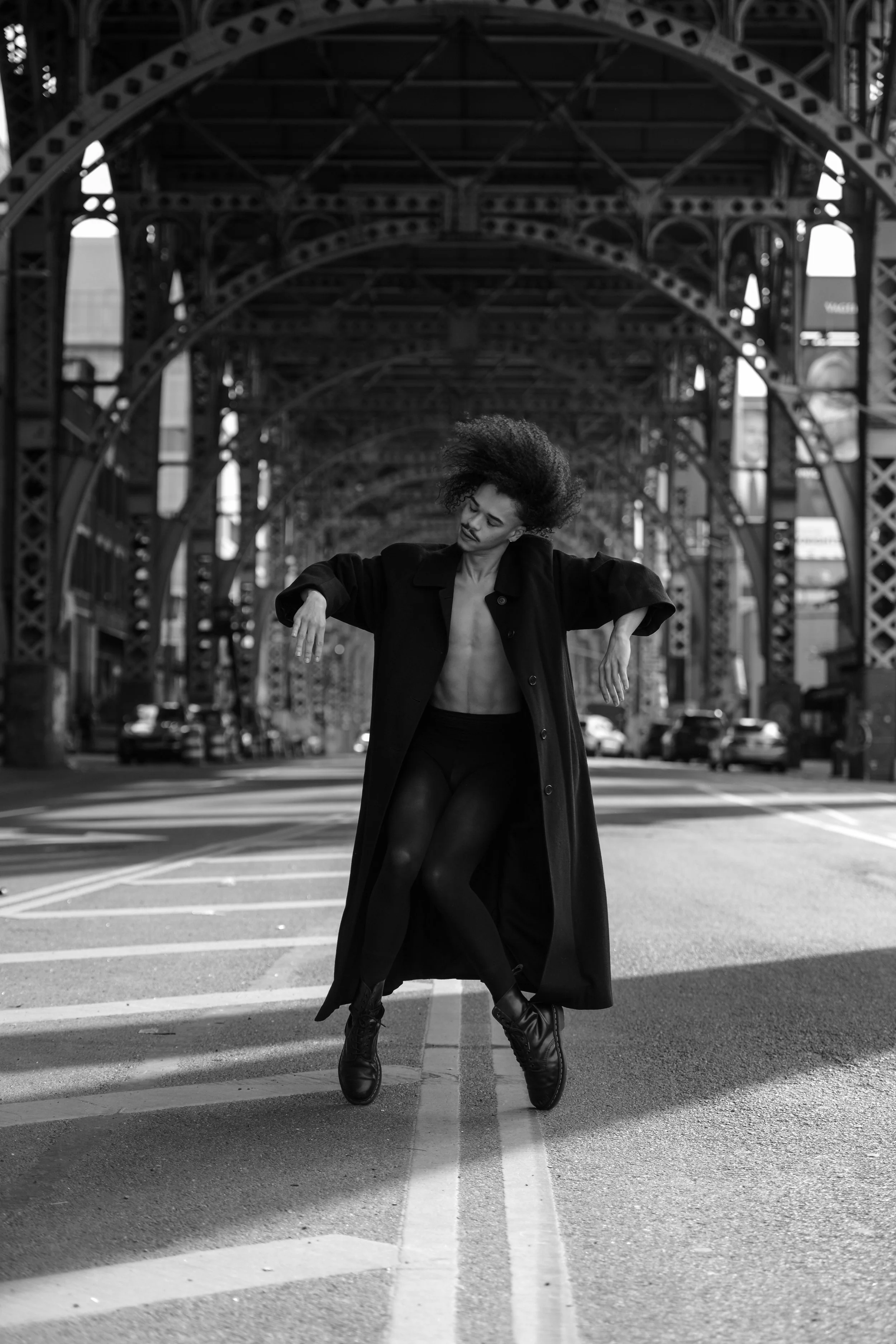 A person with curly hair dancing on a city street under an arched iron bridge, dressed in black coat, tights, and boots, captured in black and white.