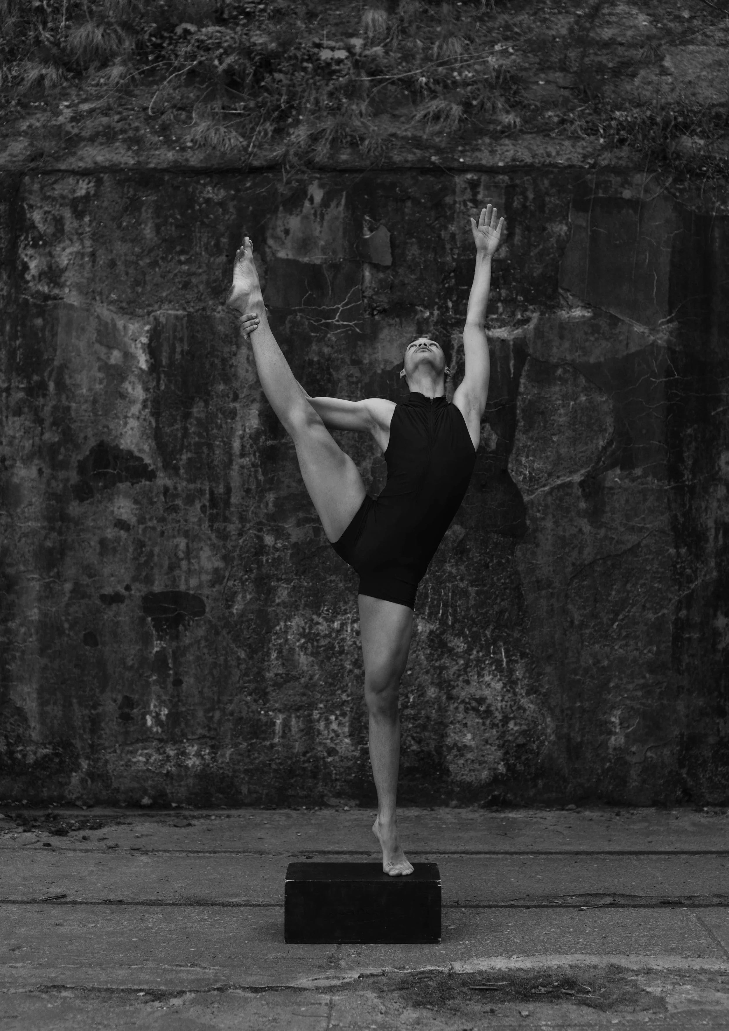 A man in a black dress performs a ballet pose on a small platform, standing on one foot with her other leg bent upward and held with her hand. She reaches upward with one arm against a rough stone wall background.