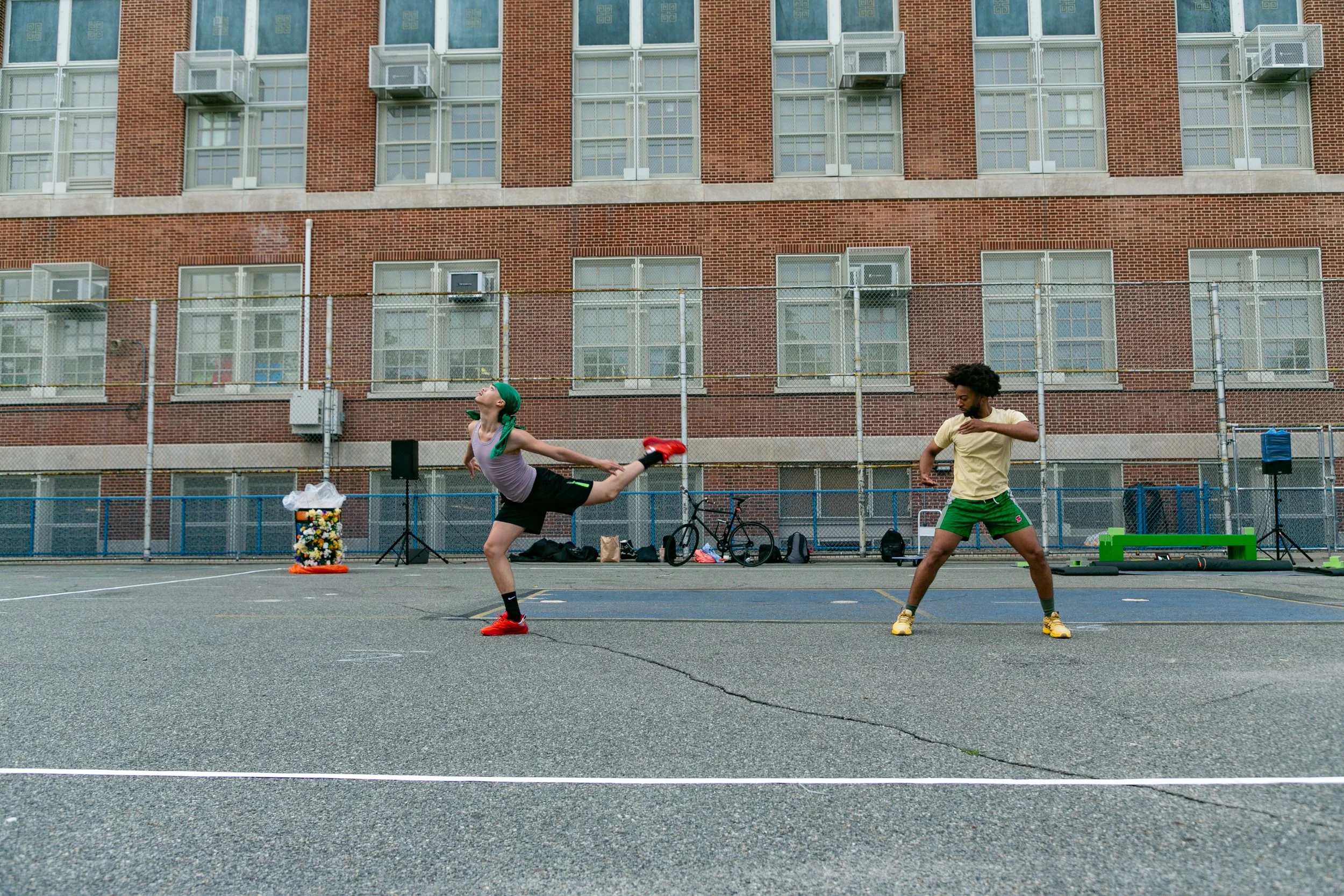 Two people exercising on an outdoor court in front of a brick building. One person is doing a kick with their right leg raised high, while the other person is standing with their arms crossed.