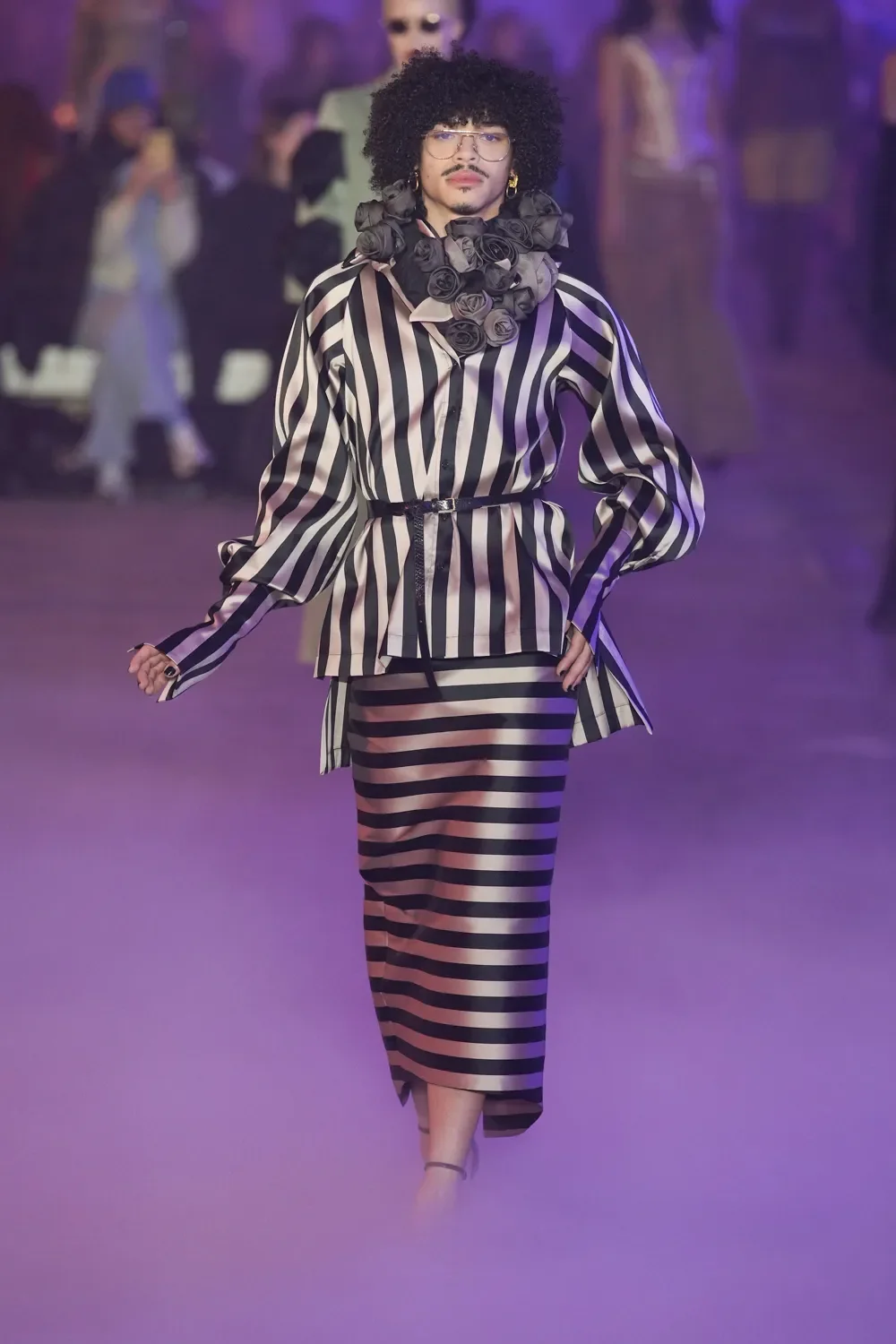 A person walking on a runway wearing a black and white striped outfit, accessorized with a large flower collar and glasses, with a purple foggy background.