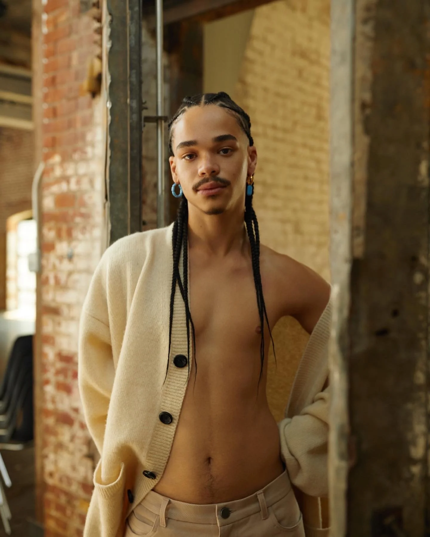 Person with braided hair wearing earrings, a beige cardigan, and beige pants, standing in front of a mirror in an industrial-style room with exposed brick walls.