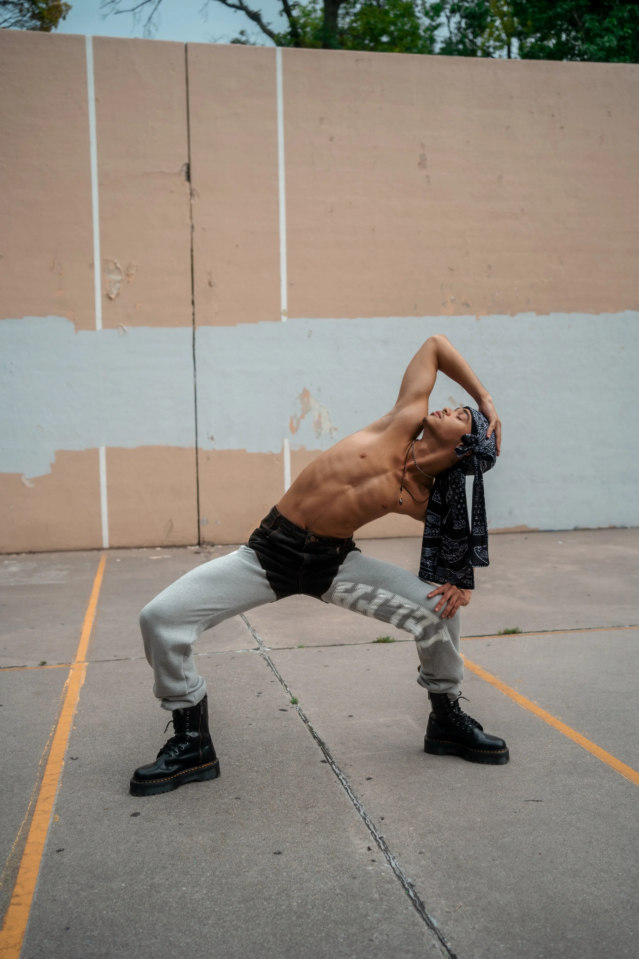 A shirtless person with a scarf on their head, wearing grey sweatpants and black boots, posing in a stretch stance on an outdoor concrete court with a painted wall and trees in the background.