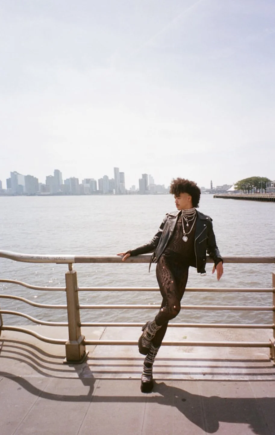 Person posing by the water on a city waterfront, wearing a black leather jacket, patterned leggings, and multiple necklaces, with a city skyline in the background.
