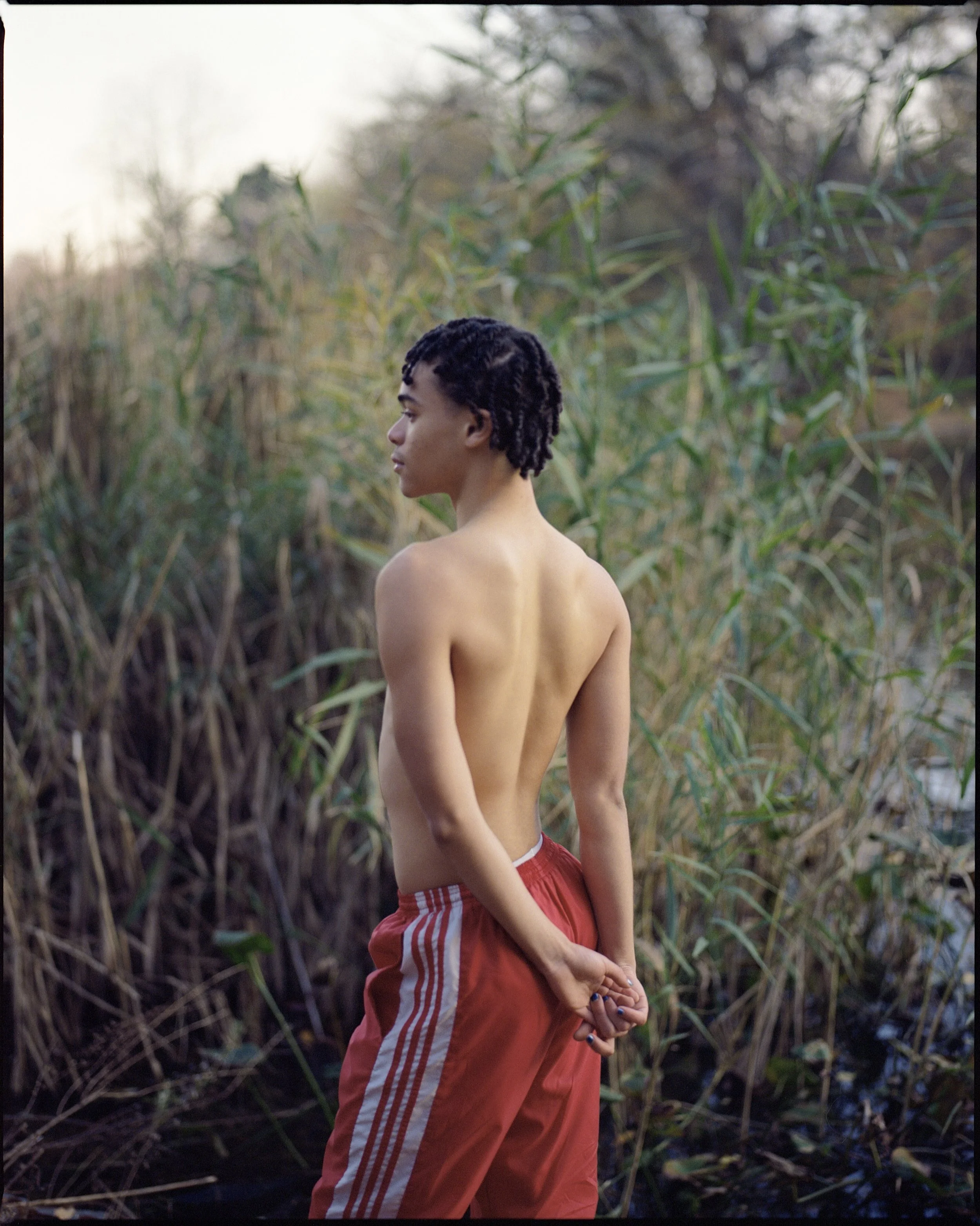 A shirtless person with short, curly hair standing outdoors near tall grass and trees, facing sideways with hands clasped behind their back.