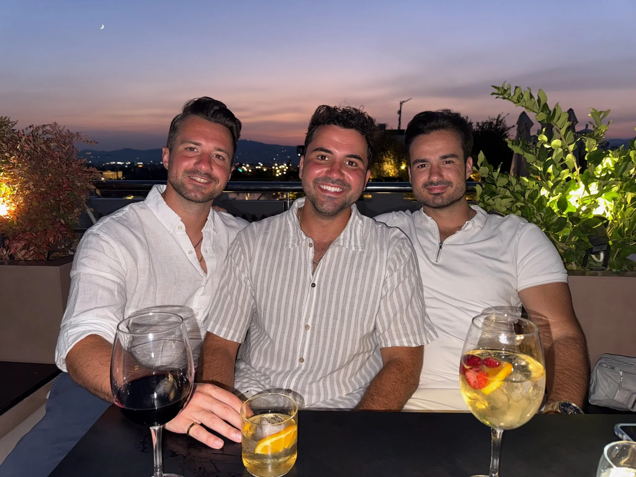 Three men smiling at a rooftop restaurant during sunset with drinks in front of them.
