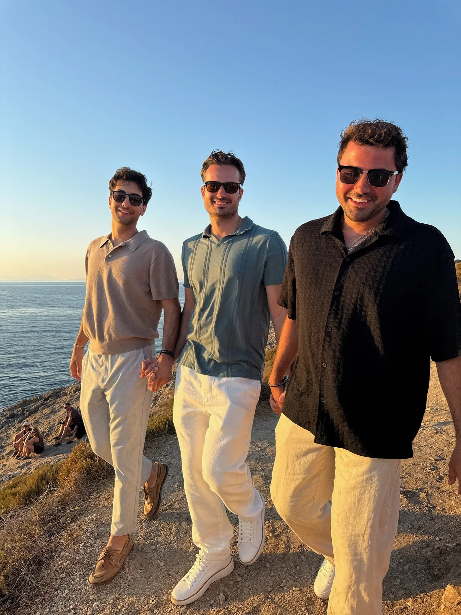 Three men walking on rocky terrain near a body of water during sunset, all wearing sunglasses and casual summer clothing.