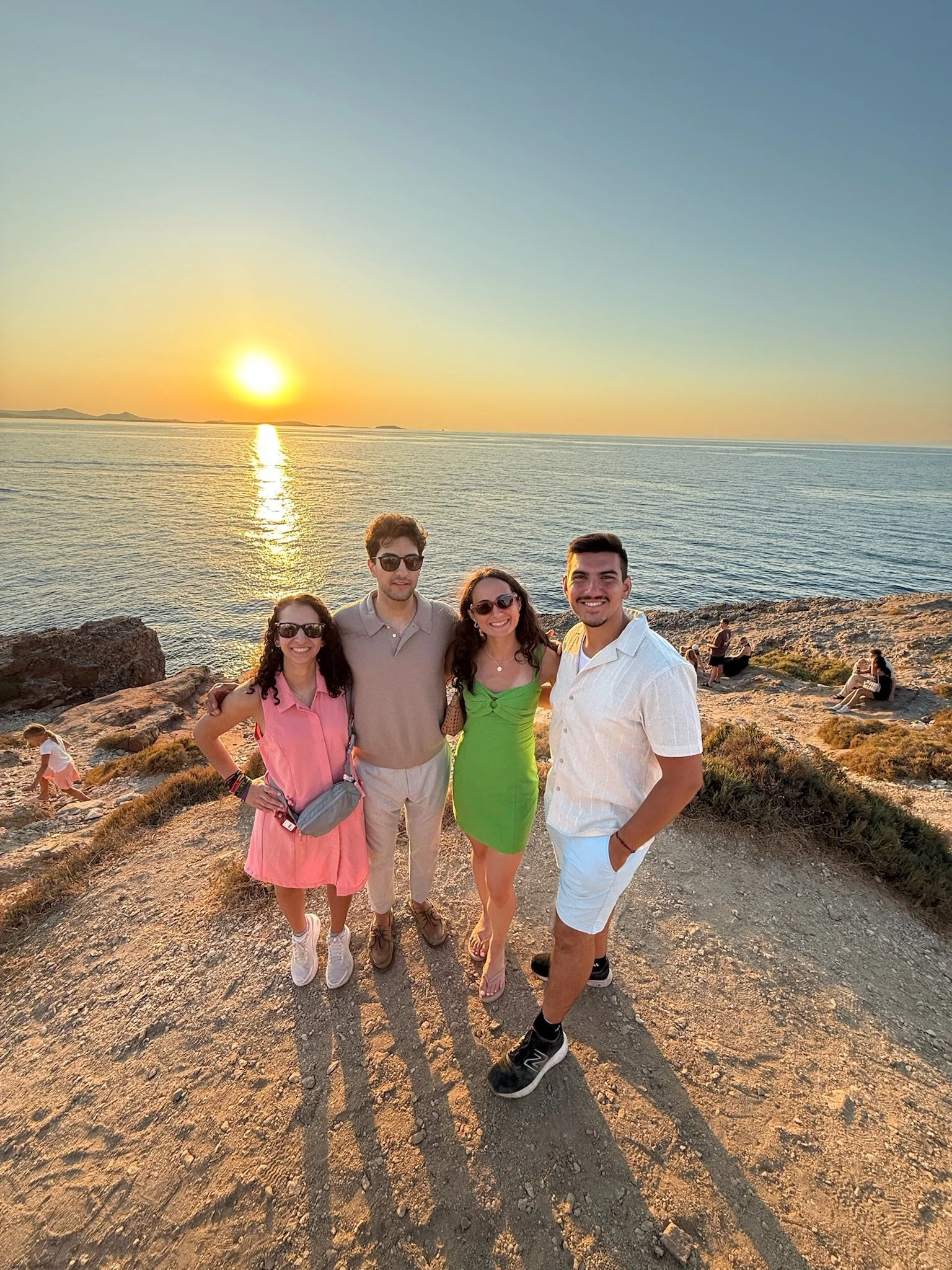 Four friends standing on a rocky coastal area at sunset, smiling for the camera with the ocean and setting sun in the background.