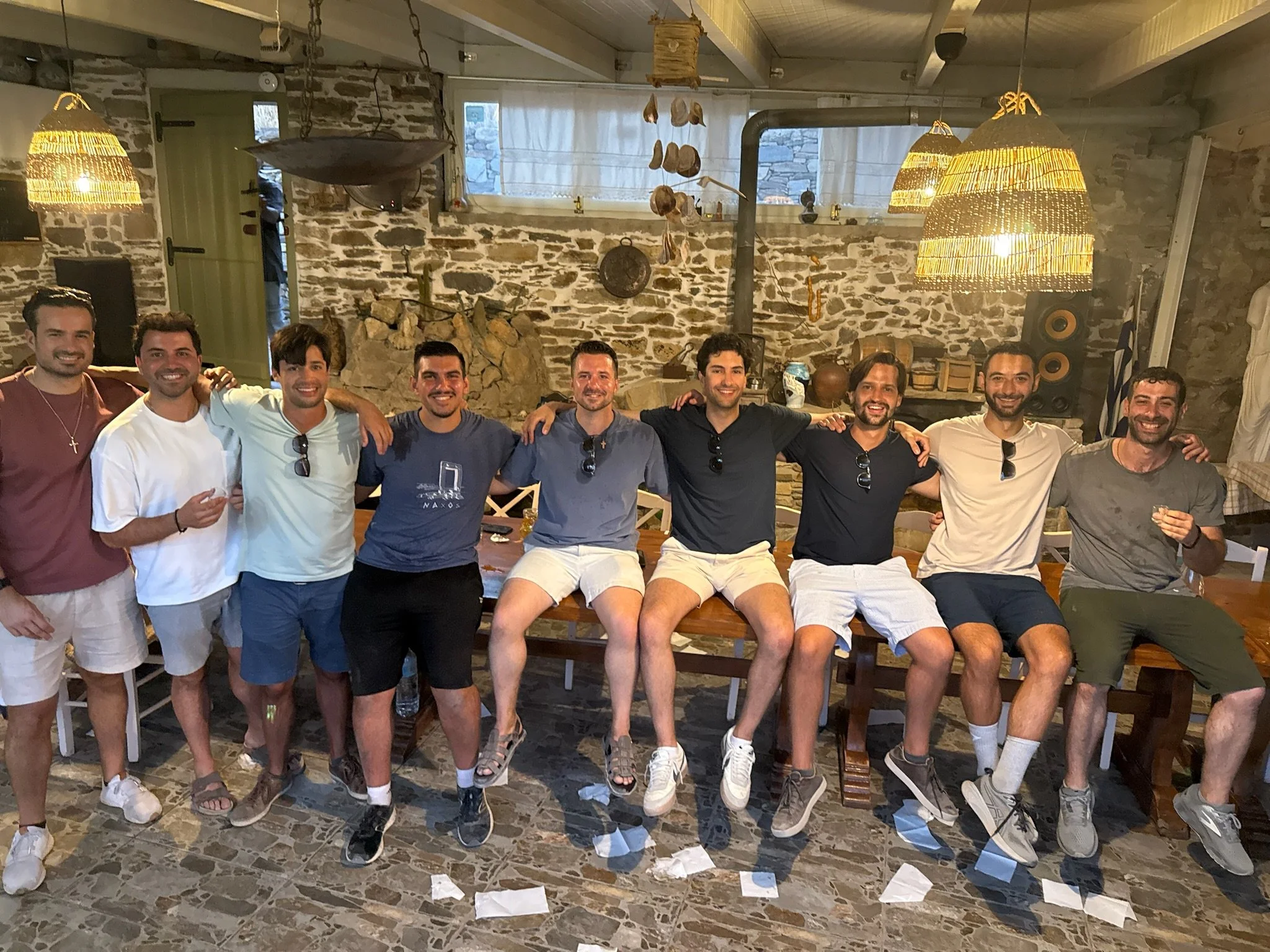 A group of nine young men sitting together on a long wooden table in a rustic room, posing for a photo with arms around each other's shoulders, smiling.