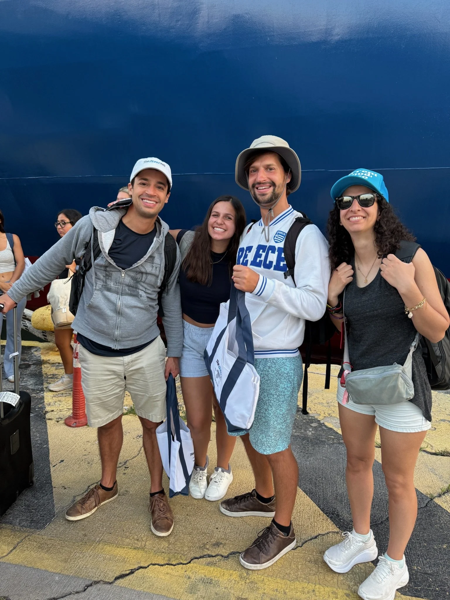 Group of five young people smiling at the airport in front of a large aircraft, carrying backpacks and tote bags, with some wearing hats and sunglasses.