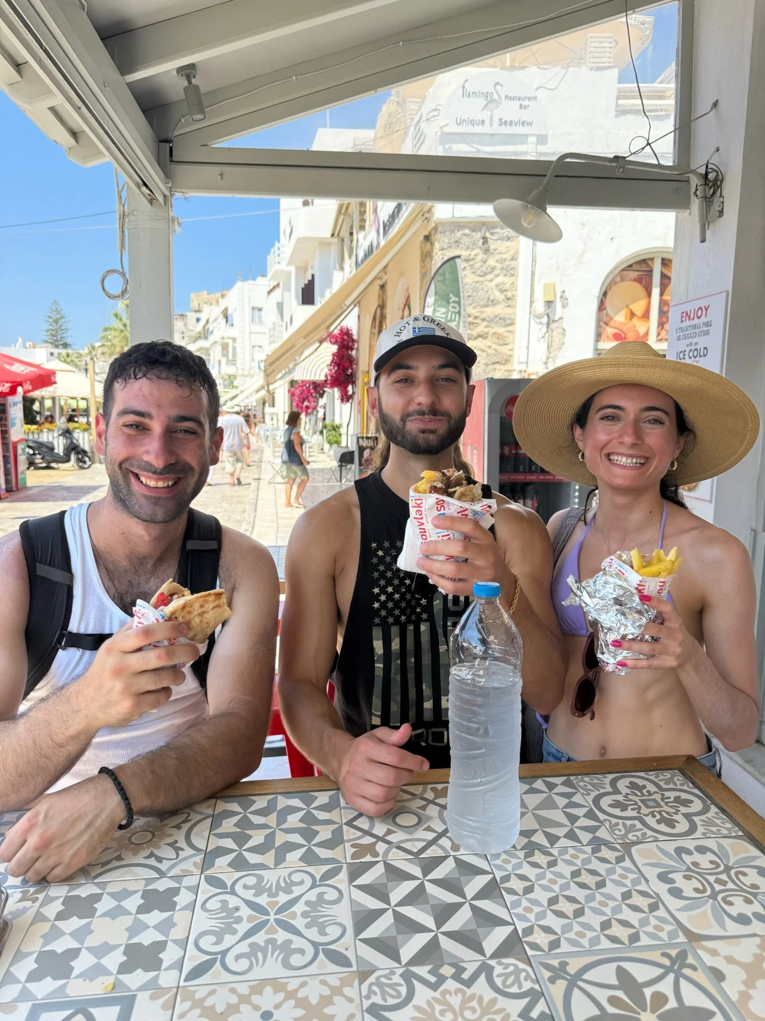 Three friends smiling and holding fries and sandwiches at an outdoor cafe, with a colorful Mediterranean-style street in the background, bright blue sky, and a water bottle on the table.