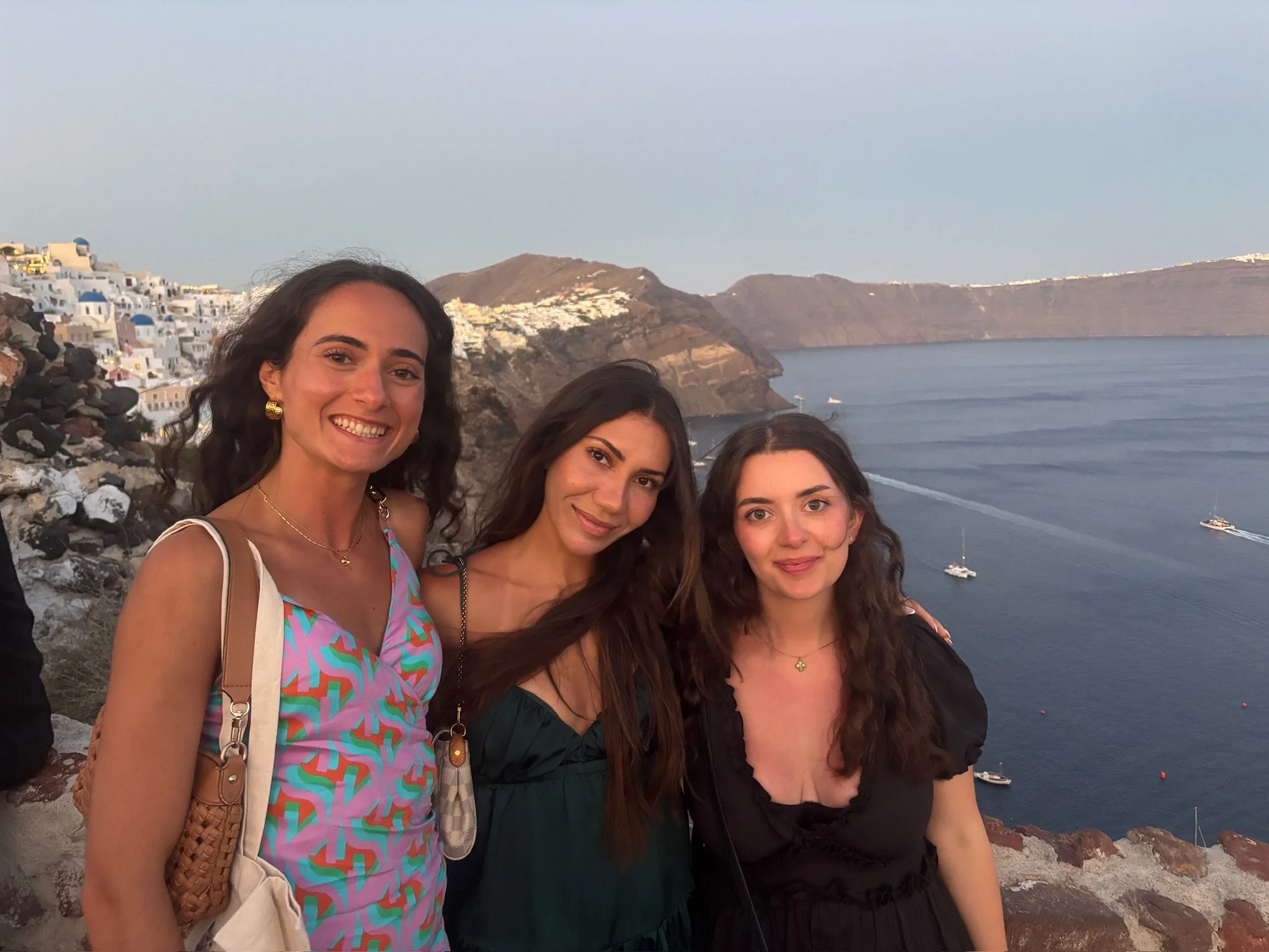 Three smiling women standing together with a scenic coastal background of mountains, white buildings, and boats on the water, during sunset.