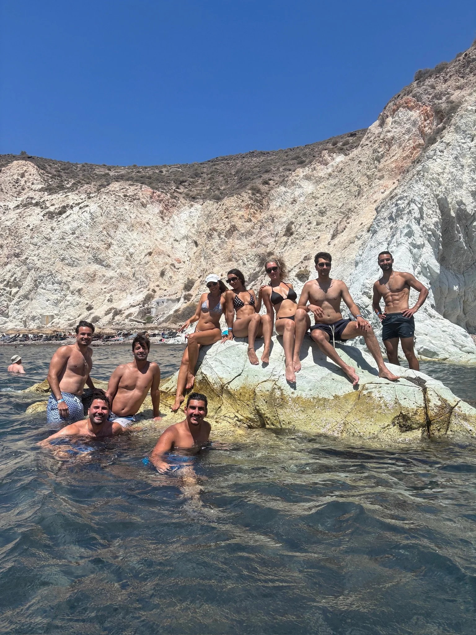 Group of eight young adults in swimwear enjoying a rocky water setting under a clear blue sky, with white cliffs in the background.