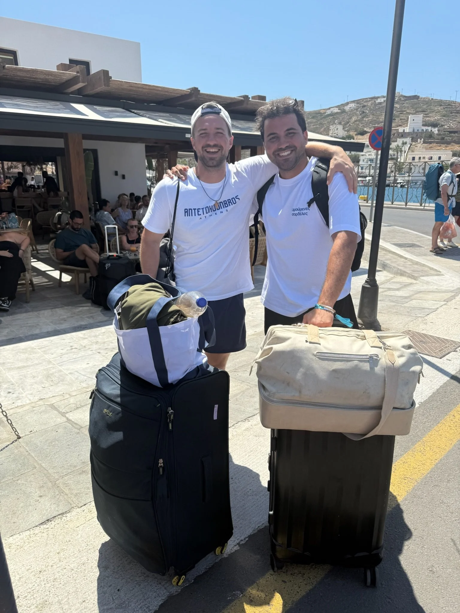 Two smiling men with backpacks and rolling suitcases standing outside near a café, with other travelers in the background.