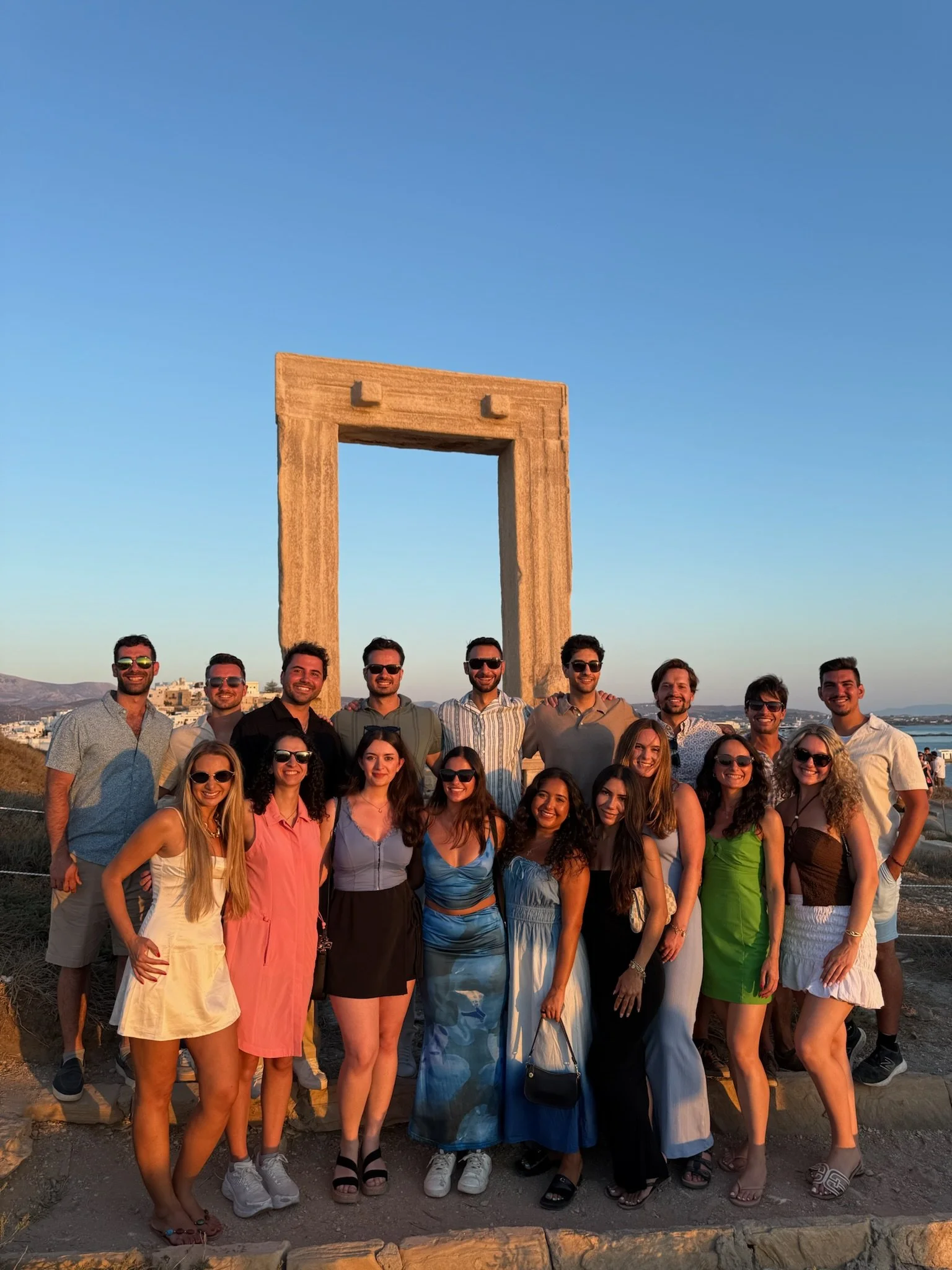 Group of 17 young adults posing outdoors at sunset in front of a large rectangular stone arch, with the ocean and distant hills in the background.