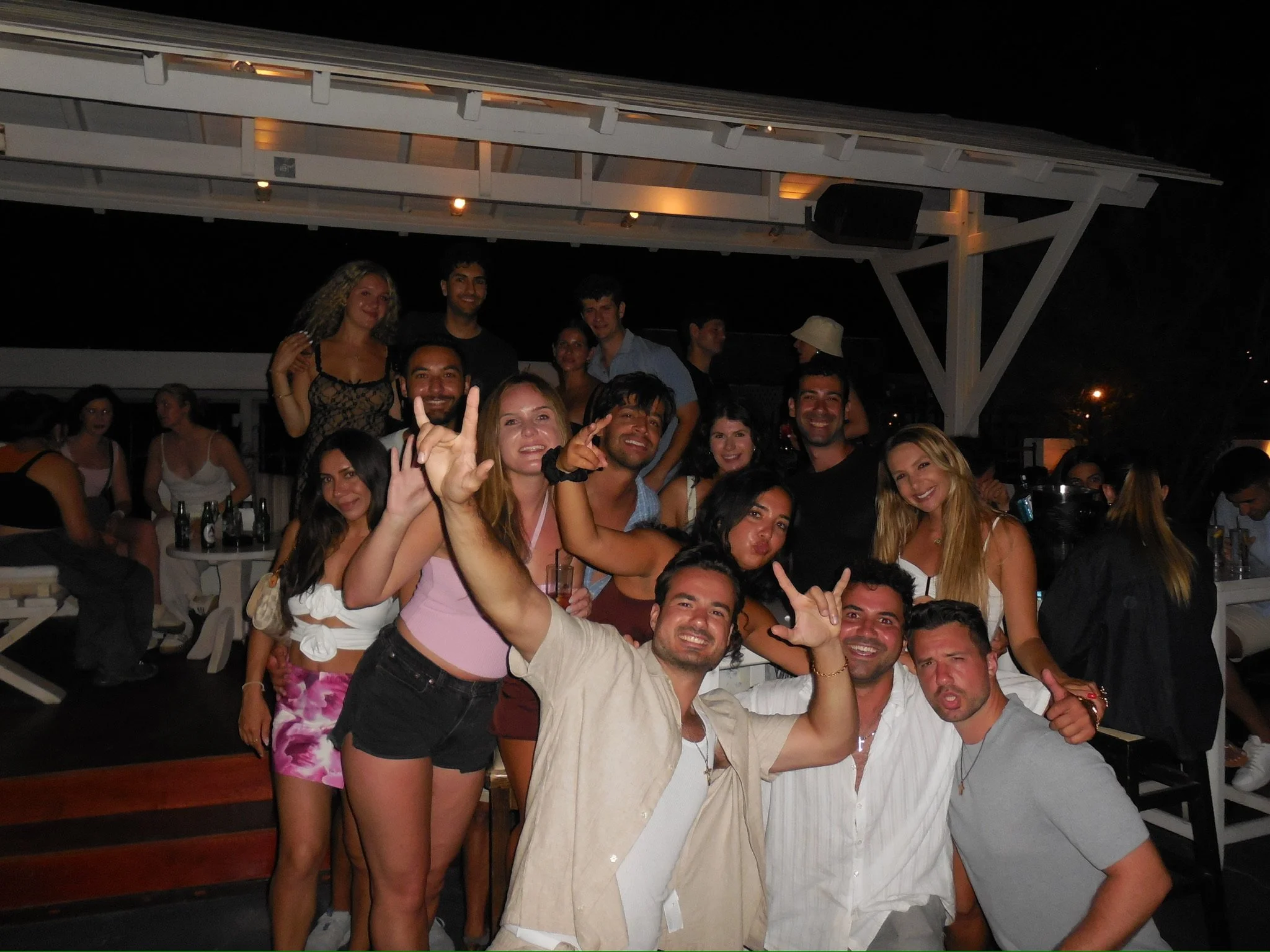 Group of young adults at a nighttime outdoor party, smiling and posing for a photo with drinks in hand, under a white wooden shelter.