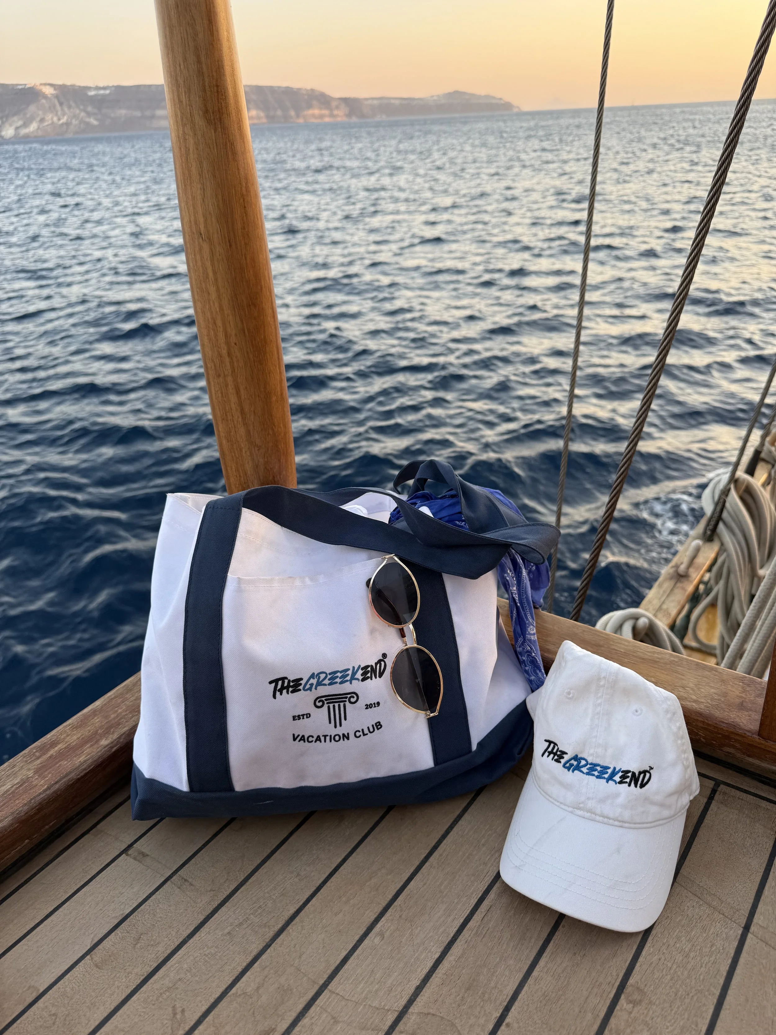 A white canvas bag with blue trim and sunglasses attached, a white cap, and a blue bandana on a boat deck with ocean and cliffs in the background at sunset.