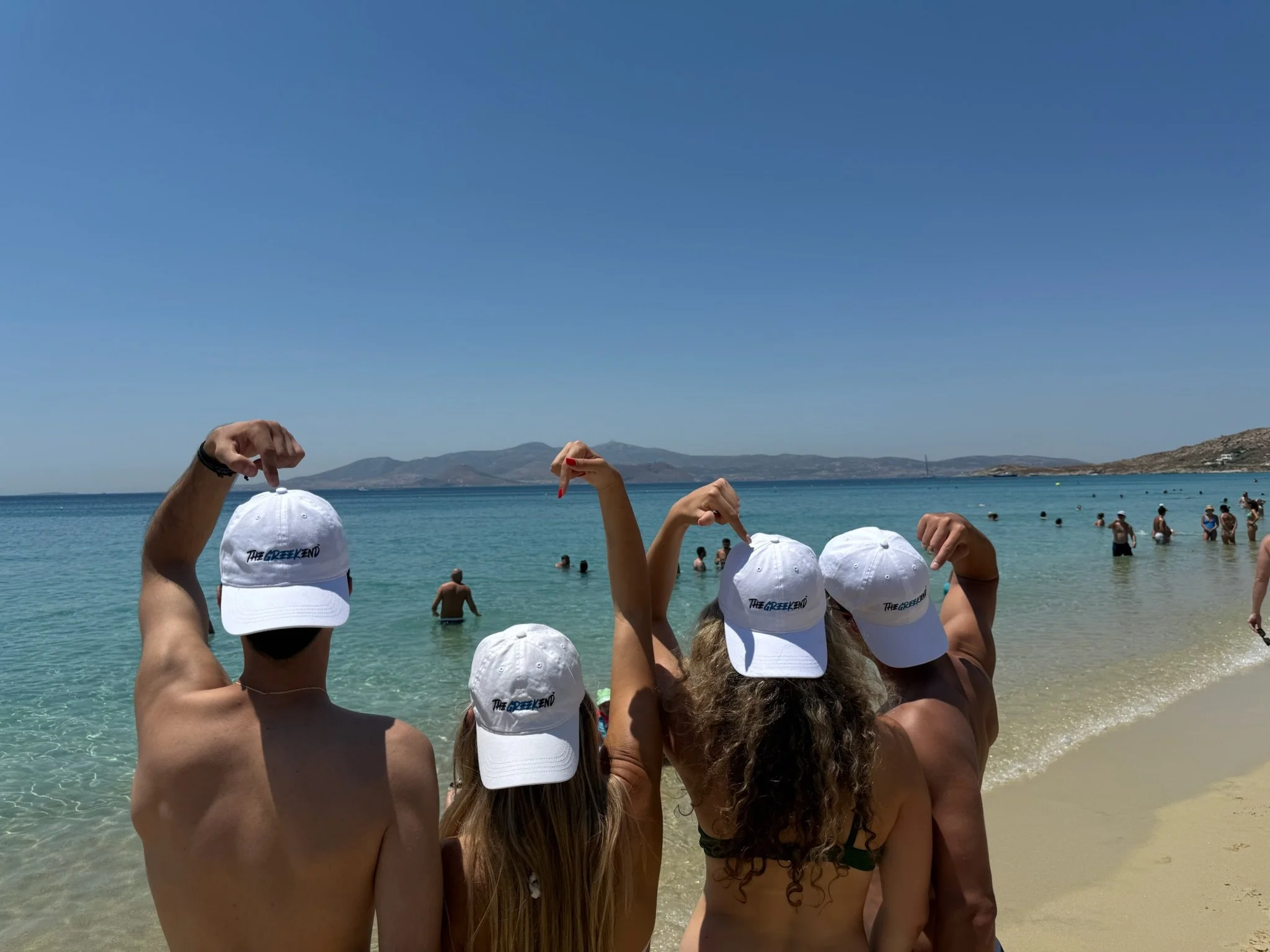 Four people at the beach wearing white hats that read 'The Greek 8ND,' posing with their back to the camera and flexing their muscles. The ocean and other beachgoers are visible in the background.