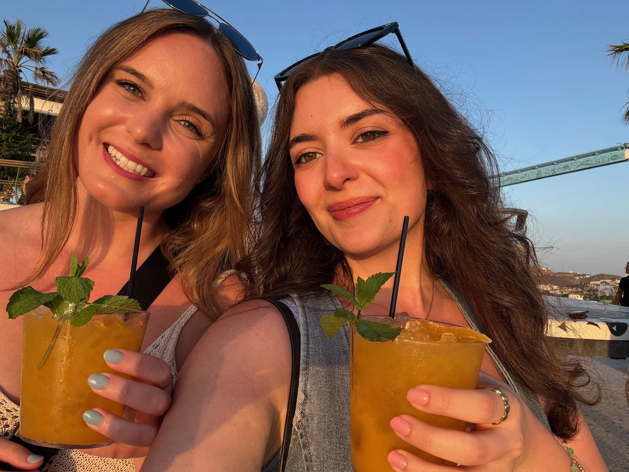 Two women smiling and holding yellow cocktails with mint leaves and straws at sunset on a beach.