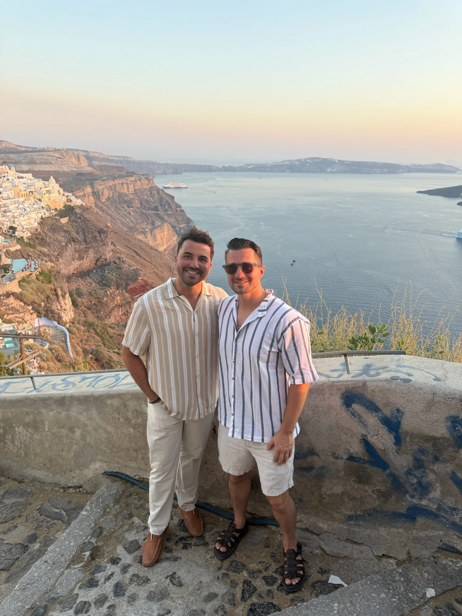 Two men standing on a stone ledge with a coastal landscape in the background, including cliffs, water, and buildings in a Mediterranean setting during sunset.