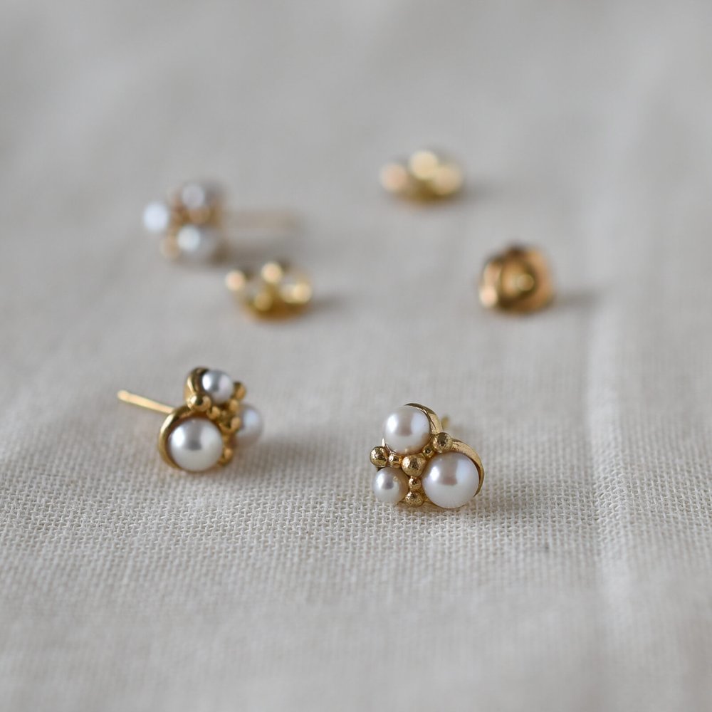 Stud earrings in gold with a cluster of three pearls of different sizes with small gold granules, shown on linen more earrings blurred in background