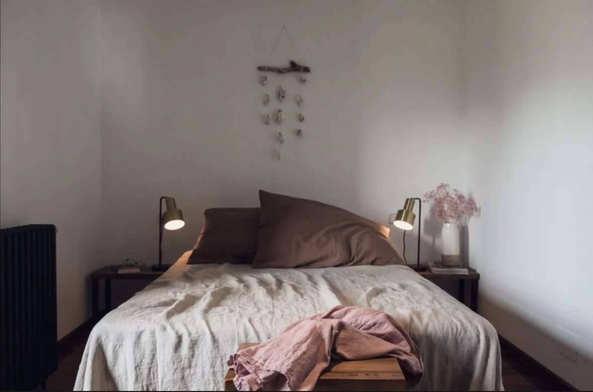 Ein gemütliches Schlafzimmer mit einem Bett, braunen Kissen und einem cremefarbenen Bettbezug. Zwei Nachttische mit Leseleuchten, ein Nachttisch mit einer Vase voller rosa Blumen, und eine Wanddekoration aus Muscheln.