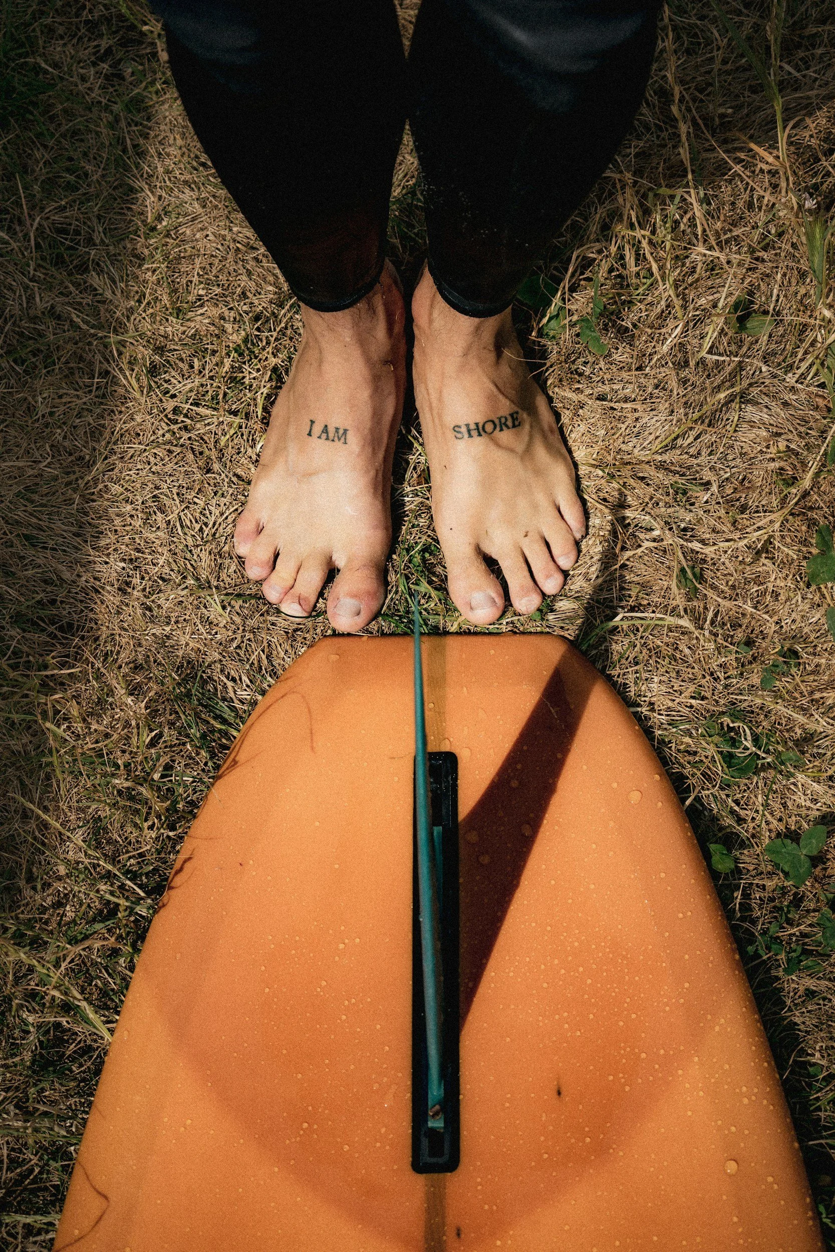 Füsse auf nassem Sand, Tattoos auf den Füßen, einer sagt "I AM" und der andere "SHORE", im Freien. Zu den Füssen liegt ein Single Fan Longboard.