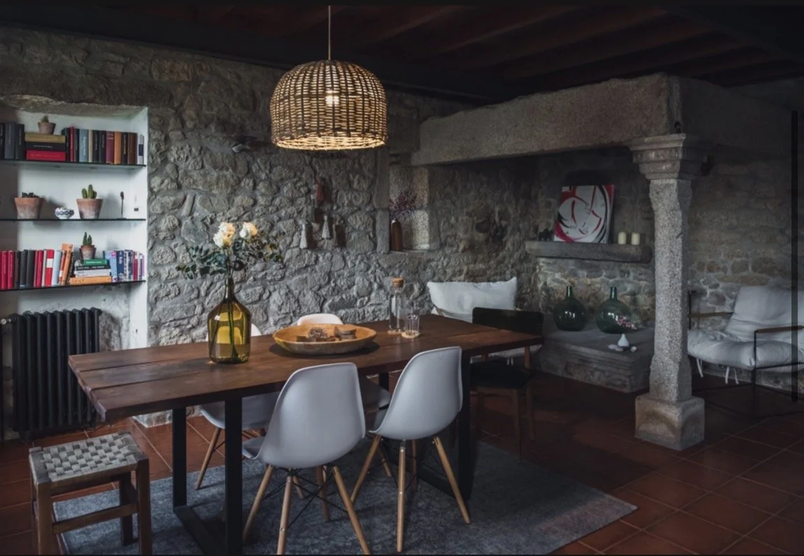 Ein rustikaler Essbereich mit einem Holztisch, weißen und schwarzen Stühlen, einer Glasvase mit Blumen, einer Wandregal mit Büchern und Pflanzen, einer Steinwand mit Kamin, Korb-Lampenleuchte, und gemütlichen Sitzgelegenheiten.