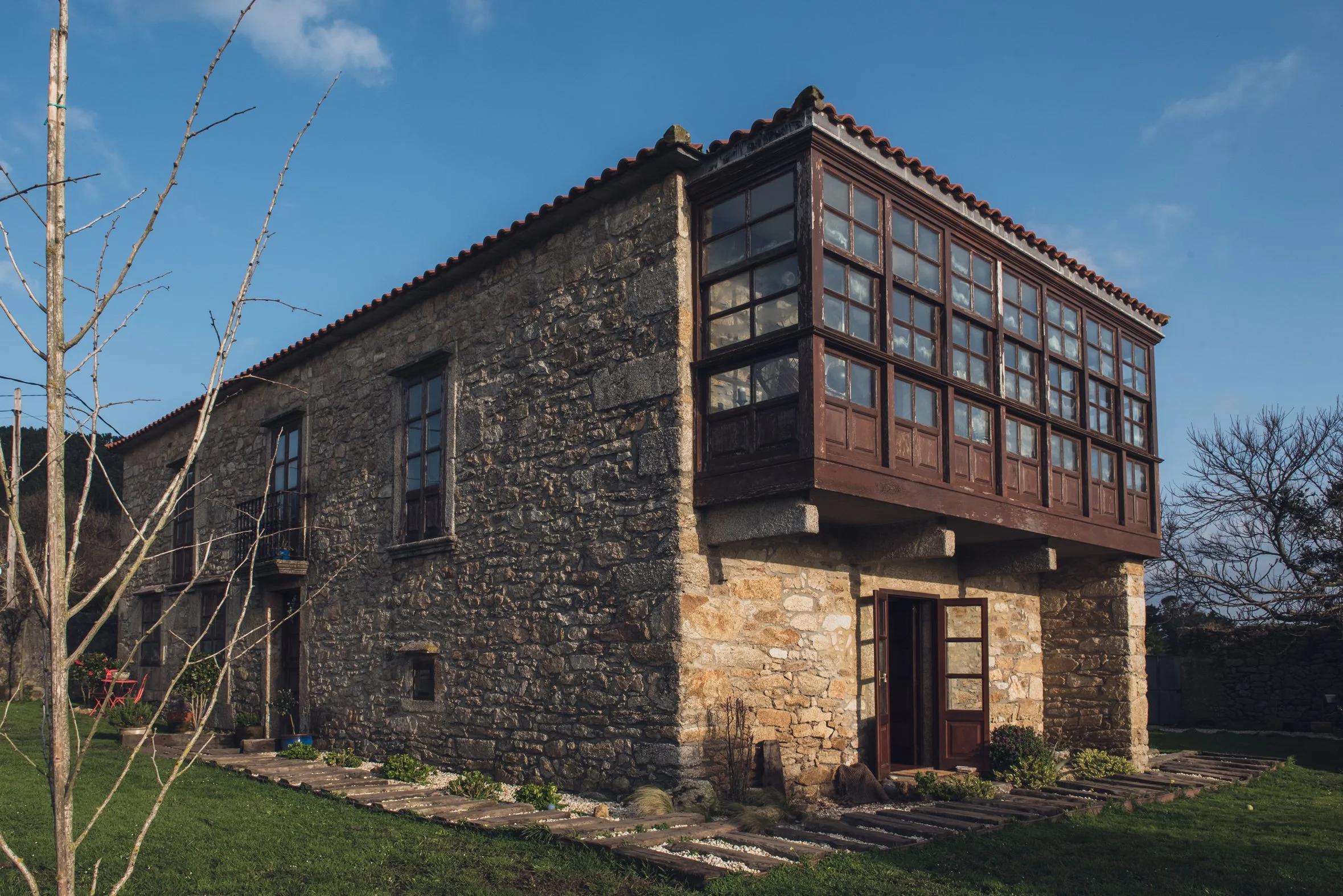 Ein altes Steinhaus mit einer Veranda an der Ecke und großen Fenstern im obersten Stock, vor einem blauen Himmel mit wenigen Wolken.