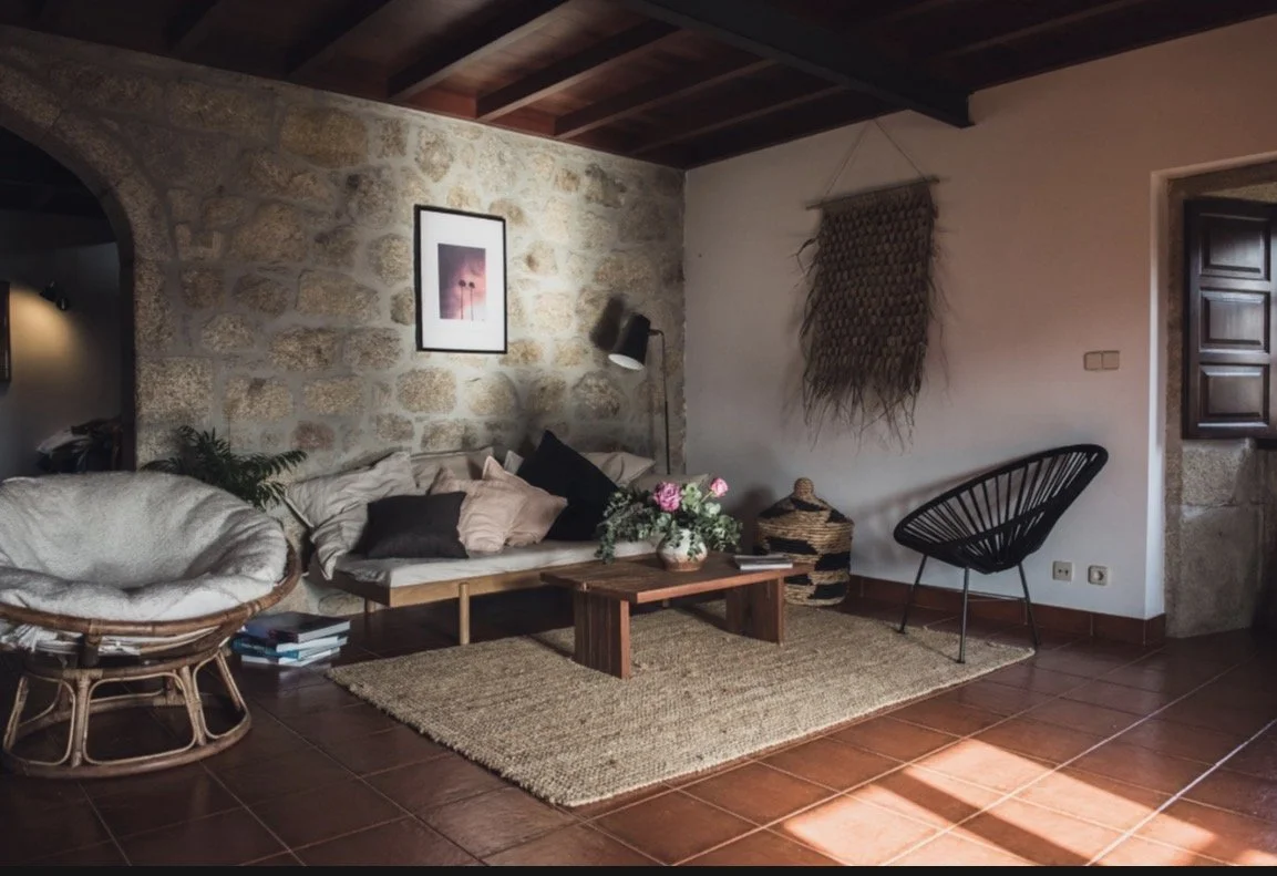 Gemütliches Wohnzimmer mit Steilwand aus Stein, Holzbalkendecke, braunem Boden, schwarzem Sessel, Holztisch mit Blumen, Wanddekoration, weichen Kissen.