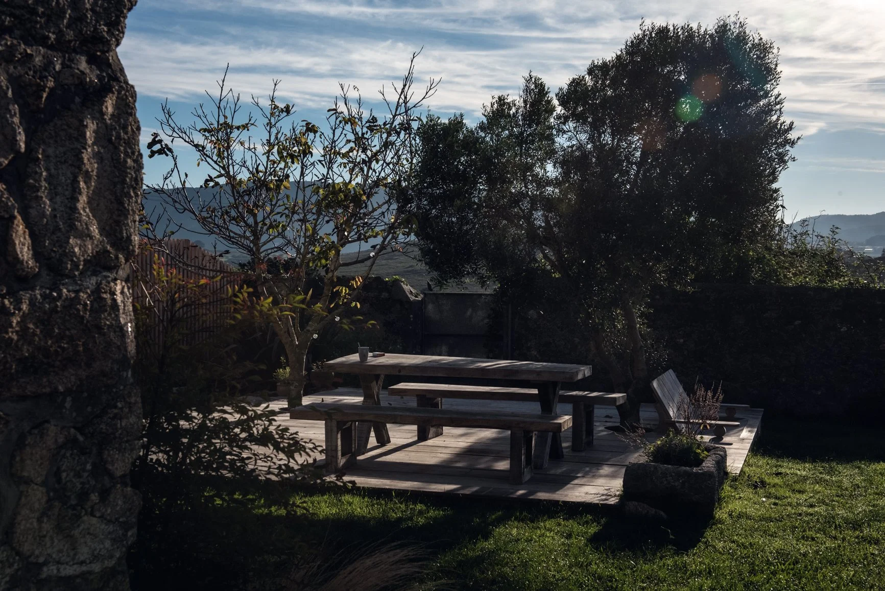 Ein Holztisch mit Sitzbänken im Garten, umgeben von Bäumen und Pflanzen, mit Bergen im Hintergrund bei Sonnenuntergang.