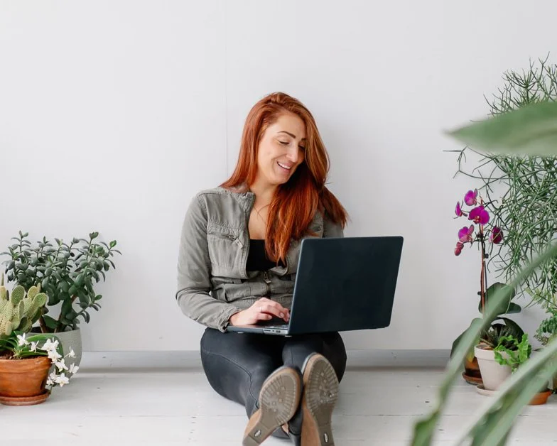Website design expert Bev Yeager sitting with potted plants around her and her computer on her lap smiling.