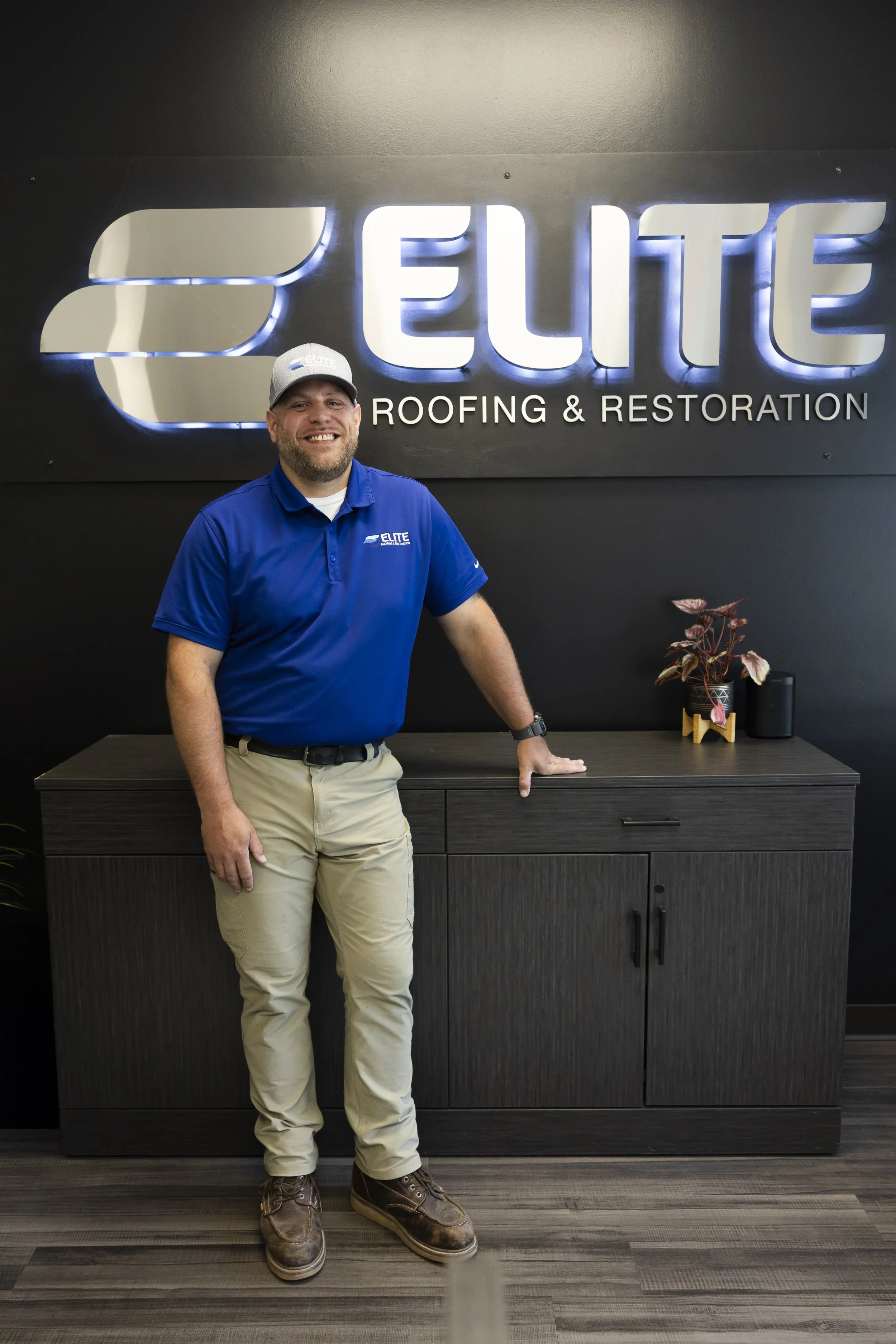 A man wearing a blue cap and shirt with 'Elite Roofing & Restoration' logos taking a selfie in front of a house while roofers work on the roof in the background.