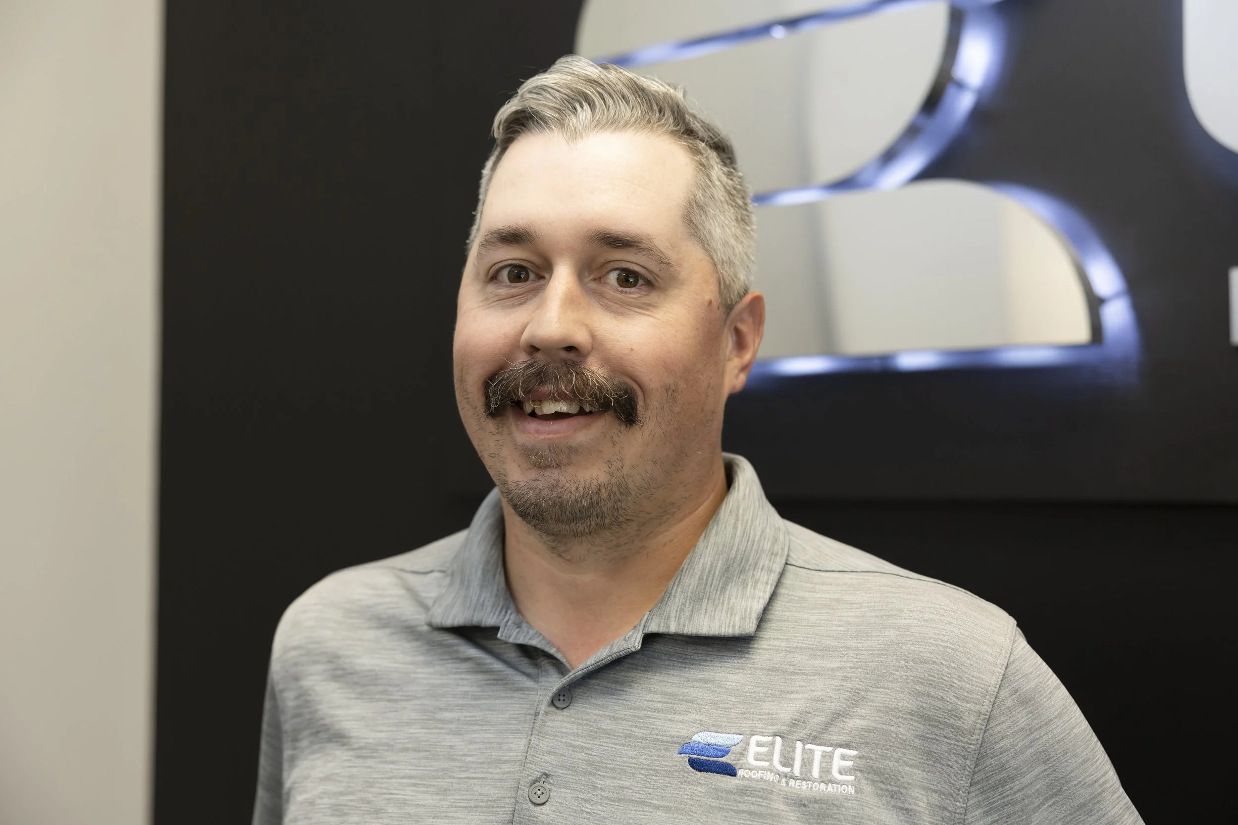 Man with a mustache and goatee wearing a gray polo shirt and a blue cap, standing in front of an American flag wall.