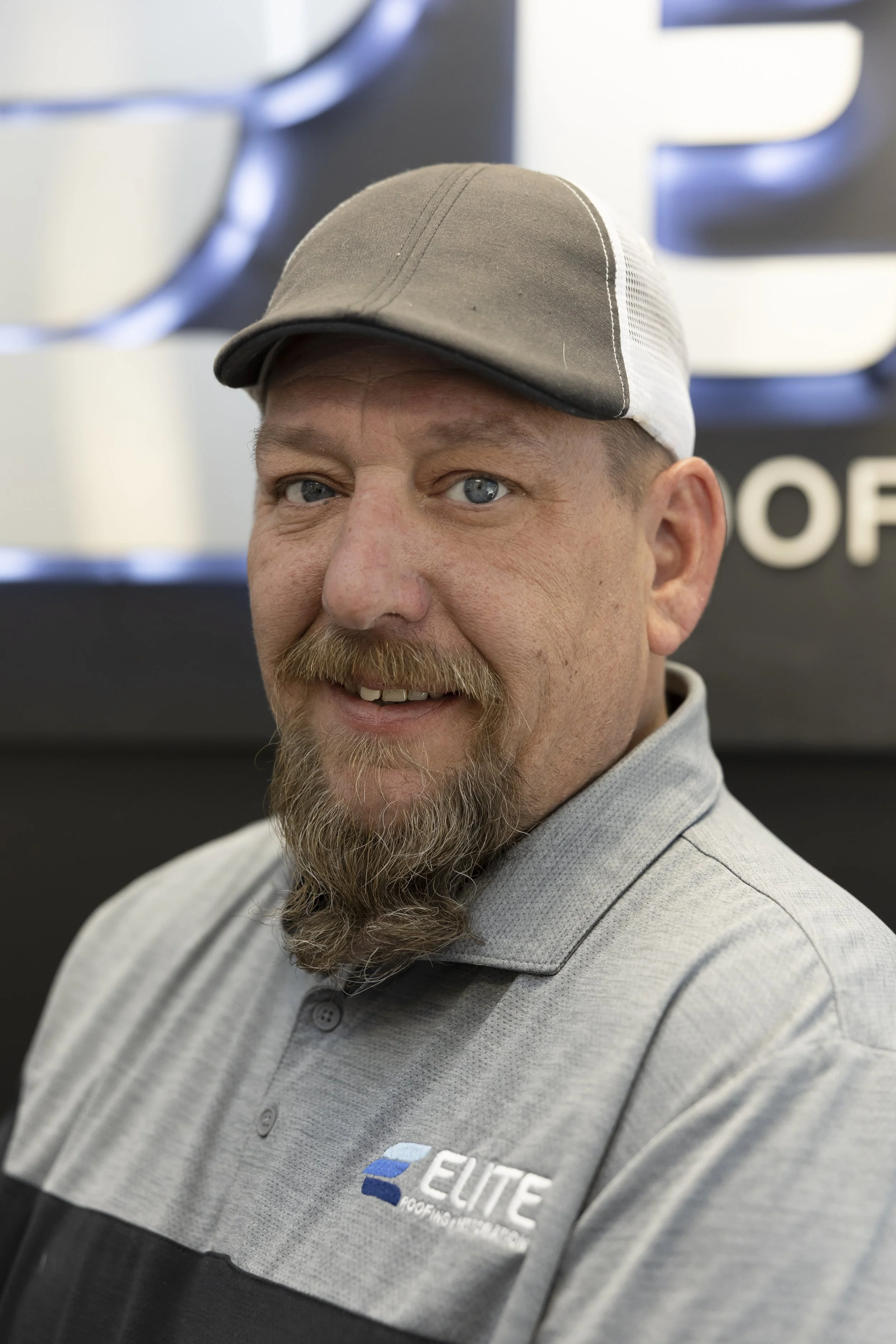 A man wearing a cowboy hat and a black jacket, smiling and standing indoors near a window.